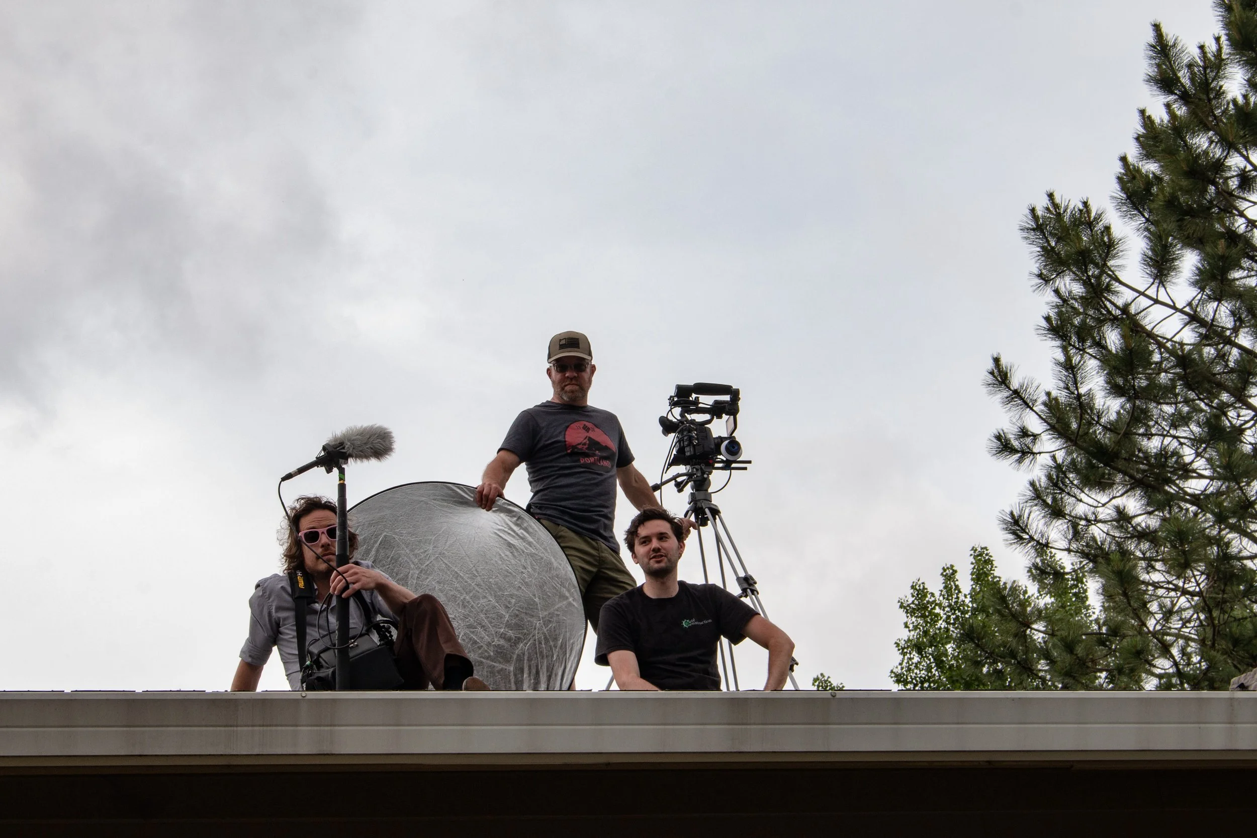 Three men on a rooftop with film equipment, two are sitting and one is standing, with trees and a cloudy sky in the background.