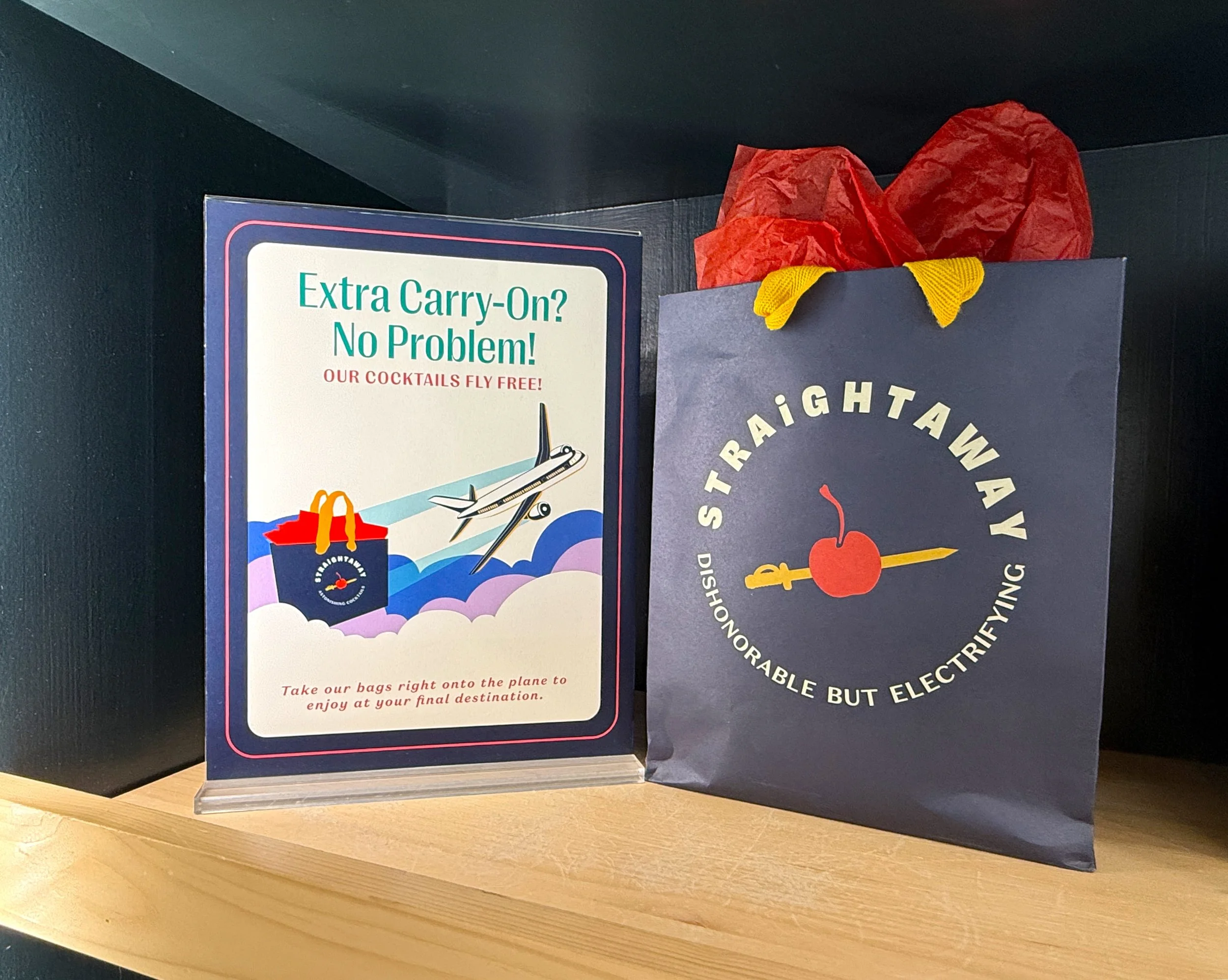 A promotional display featuring a sign about carry-on bags and a dark blue paper bag with a red cherry and yellow fork design from Straightaway. The sign says 'Extra Carry-On? No Problem! Our Cocktails Fly Free!' and mentions taking bags onto the plane for final destination enjoyment.
