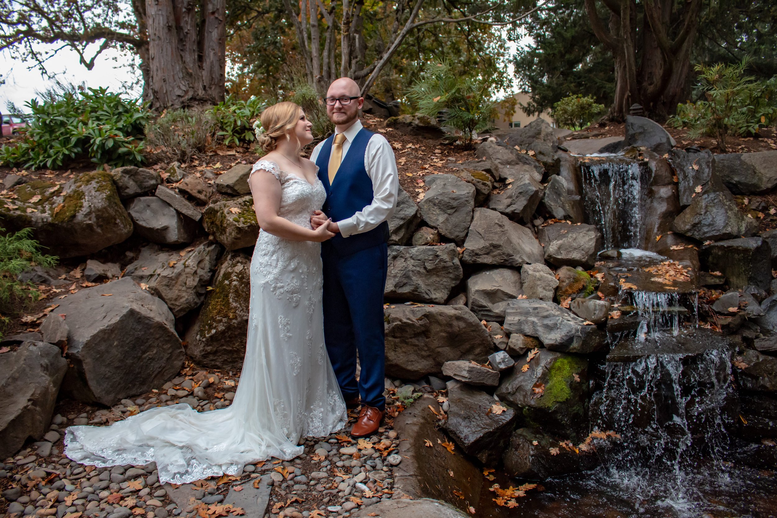 A bride and groom in wedding attire standing outdoors near a small waterfall surrounded by rocks and trees.