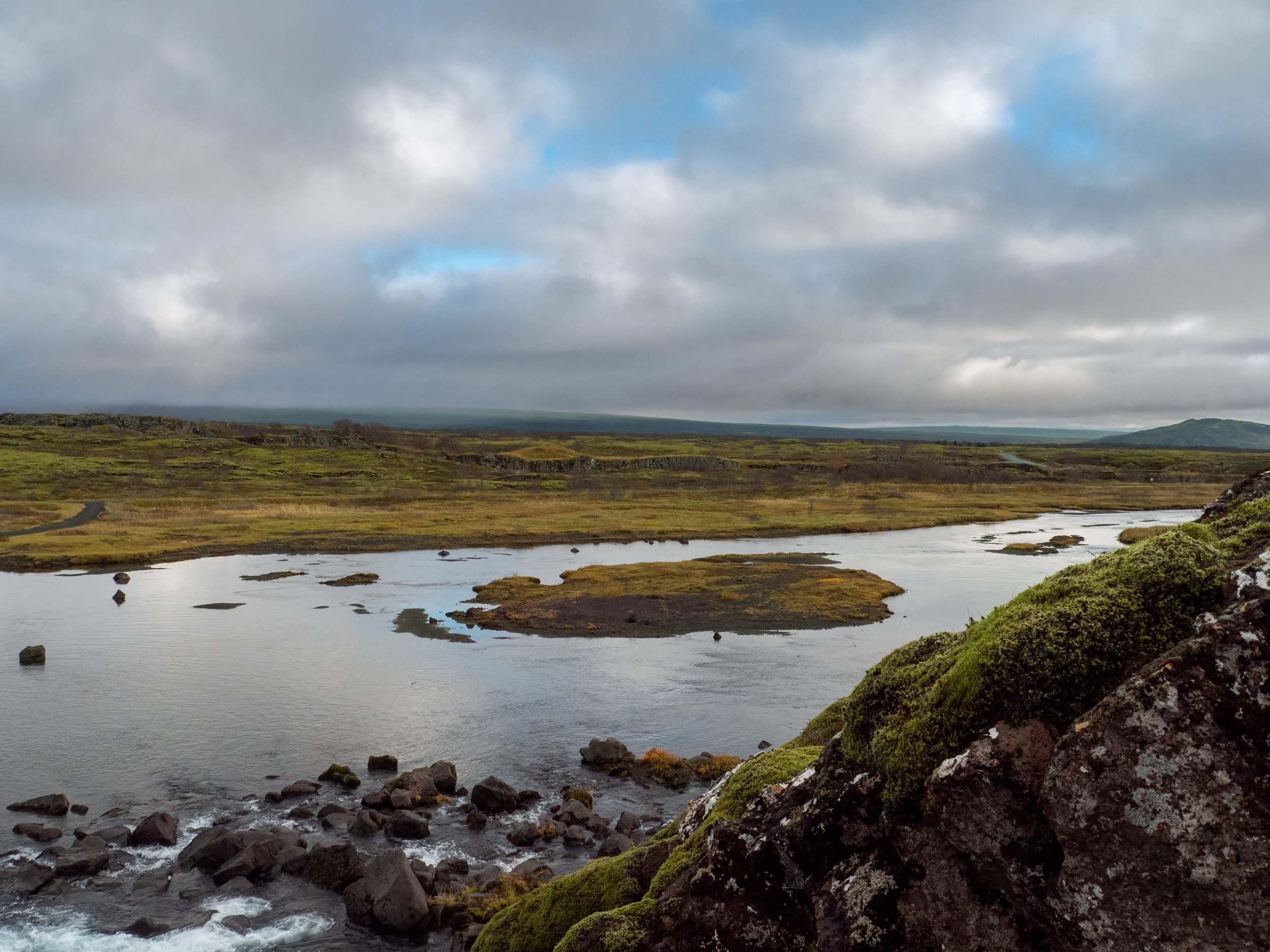 A wide river flowing through a grassy landscape with moss-covered rocks in the foreground and cloudy sky above.