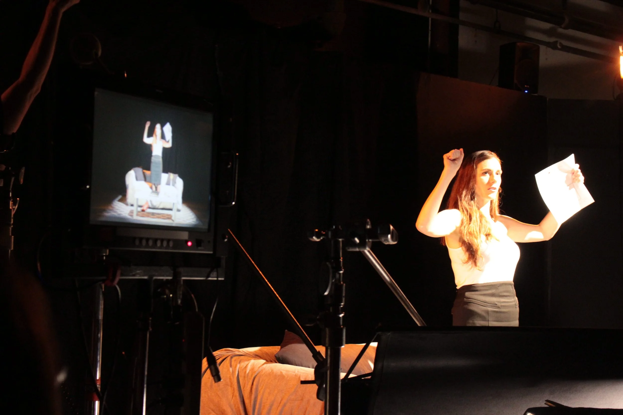 A woman on stage lifting her right fist and holding a paper in her left hand, with a monitor showing a scene of her standing on a couch with her hands raised behind her.