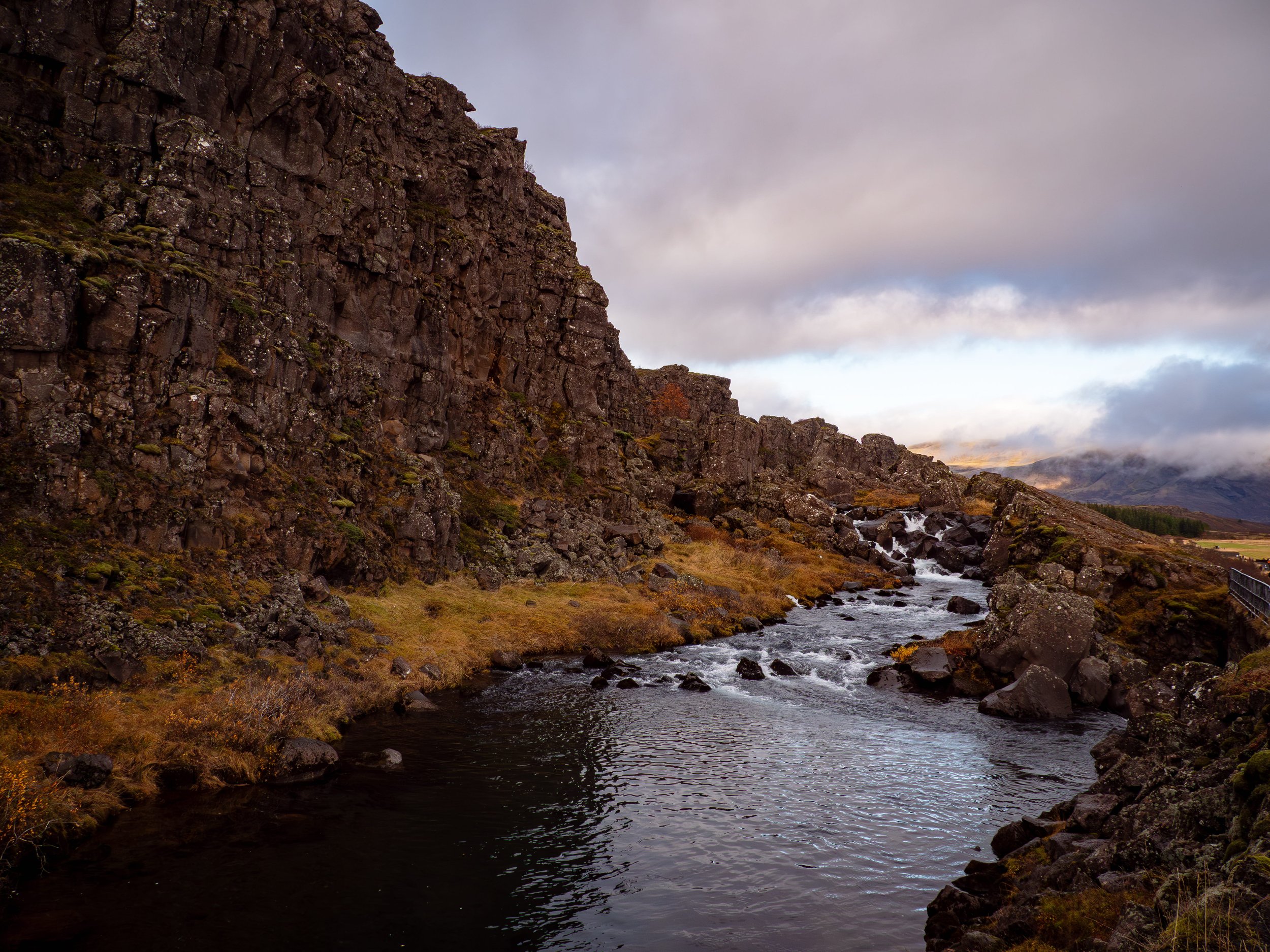 A river flowing through a rugged canyon with steep, rocky cliffs on the left and misty mountains in the distance under a cloudy sky.
