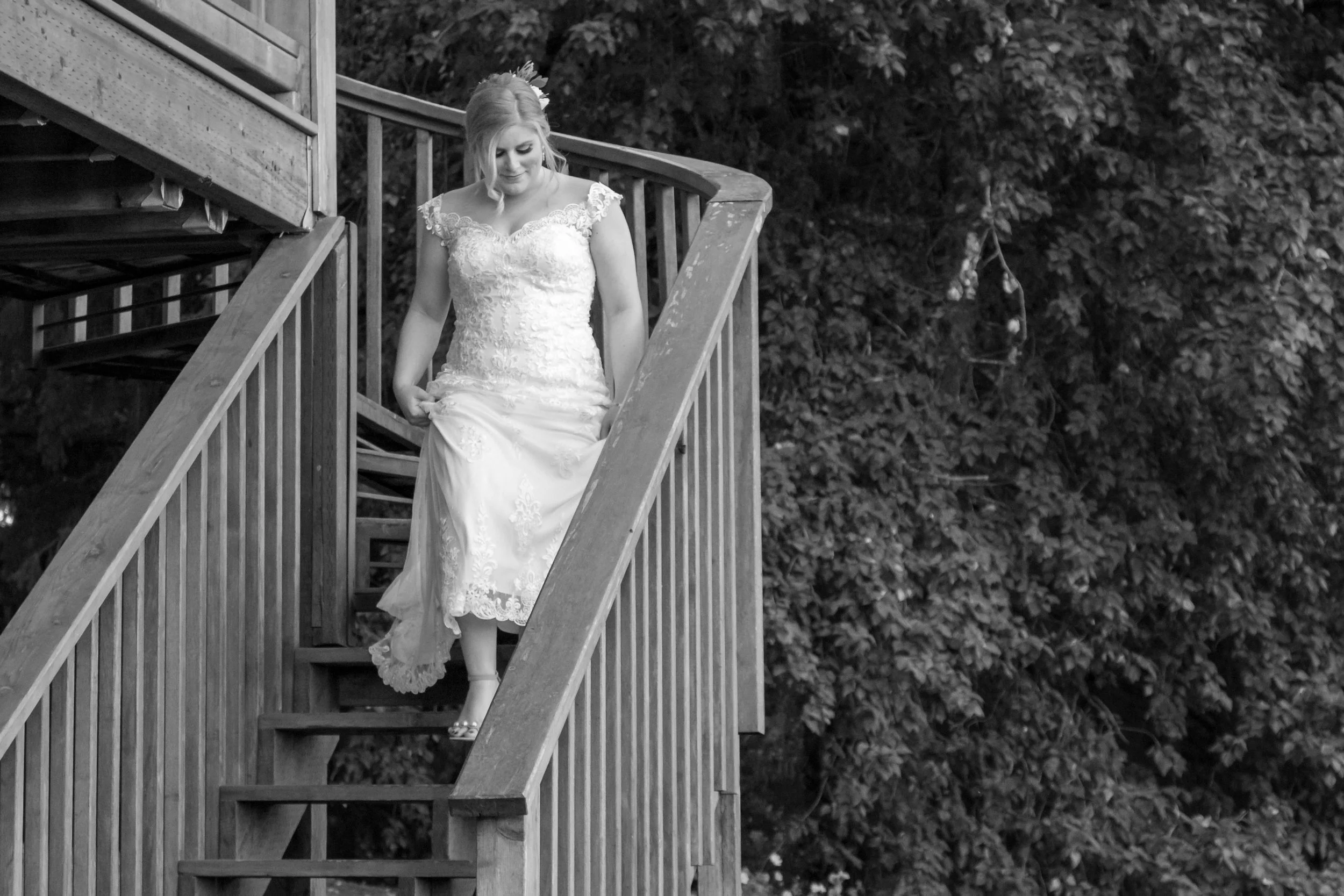 A woman in a lace wedding dress descending a wooden staircase outdoors with trees in the background.