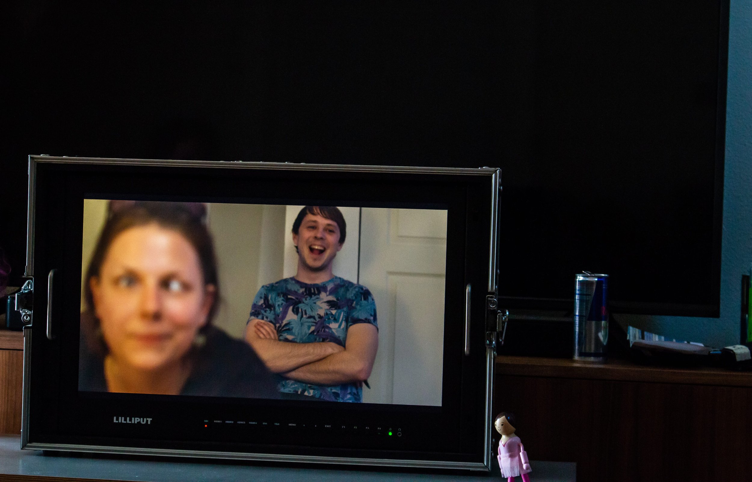 Television screen showing a blurred close-up of a woman and a clear shot of a smiling man with crossed arms. The TV stands on a wooden surface with a small doll in front.