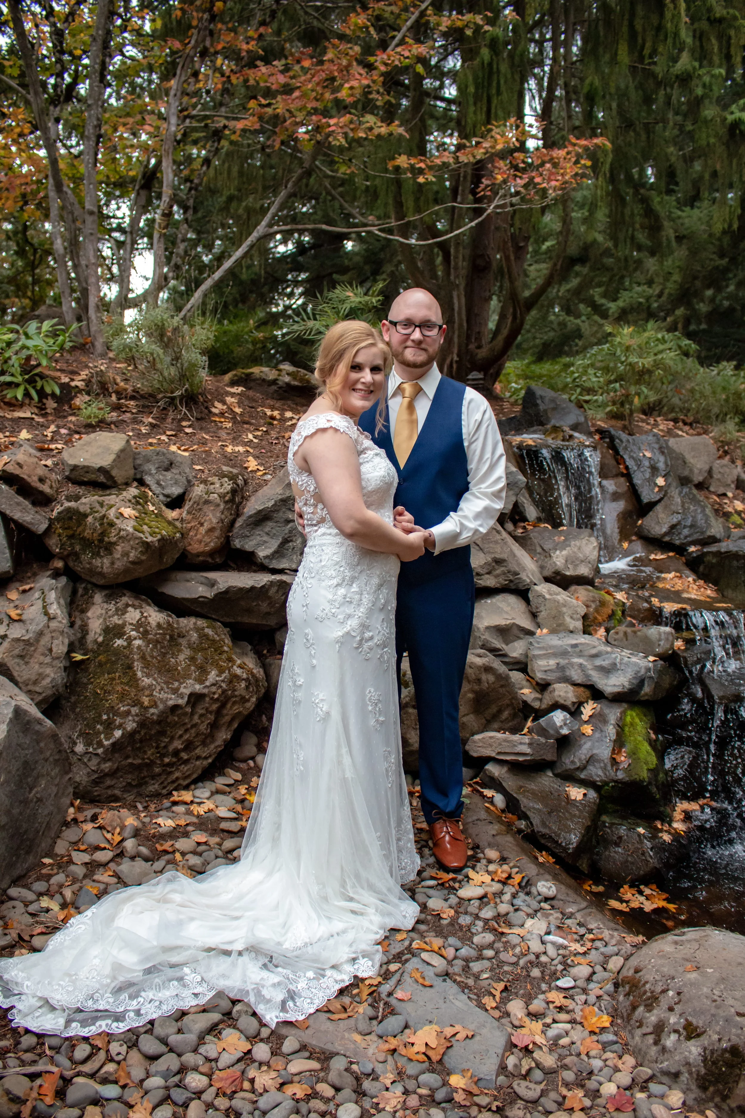 A bride and groom standing together outdoors in front of a small waterfall and rocks with autumn leaves, smiling and holding hands.