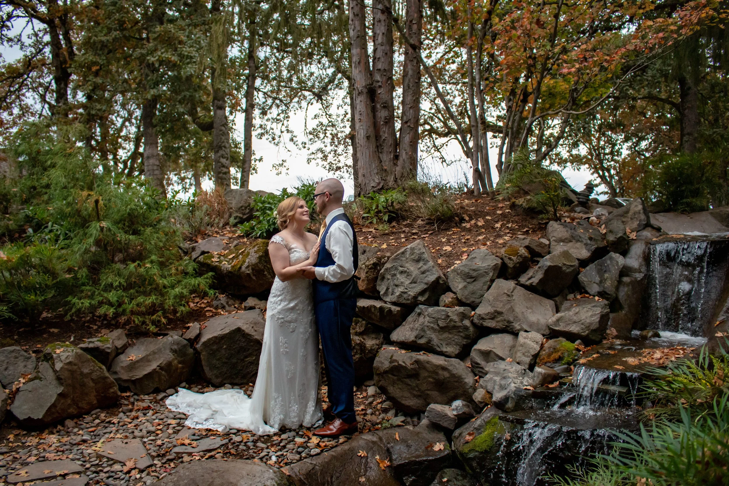 A bride and groom embrace by a small waterfall in a wooded area with trees and rocks, during their wedding ceremony.