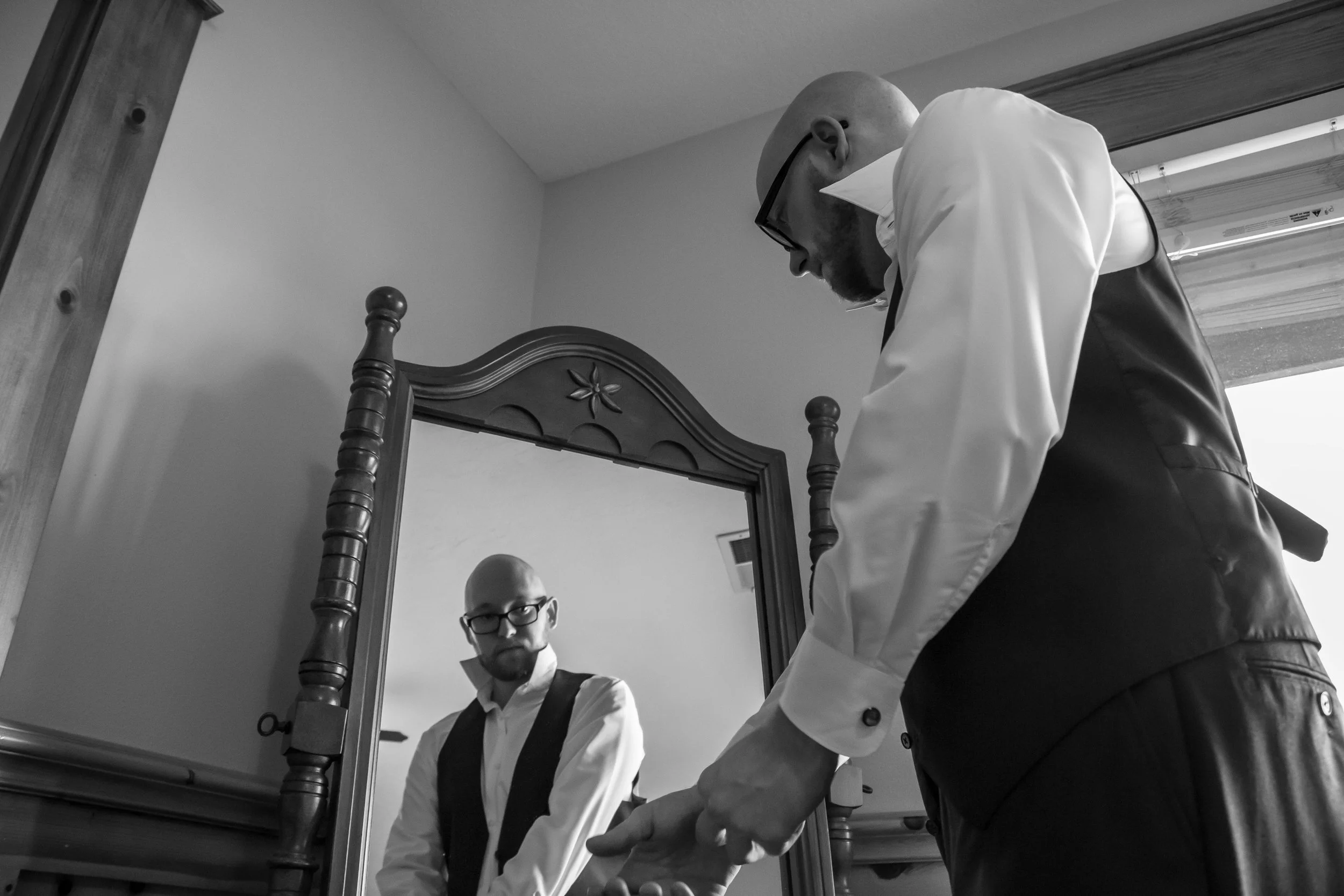A man in glasses and a vest looks into a mirror, while another man adjusts his sleeve, both dressed in formal attire, in a room with a wooden headboard and ceiling window.
