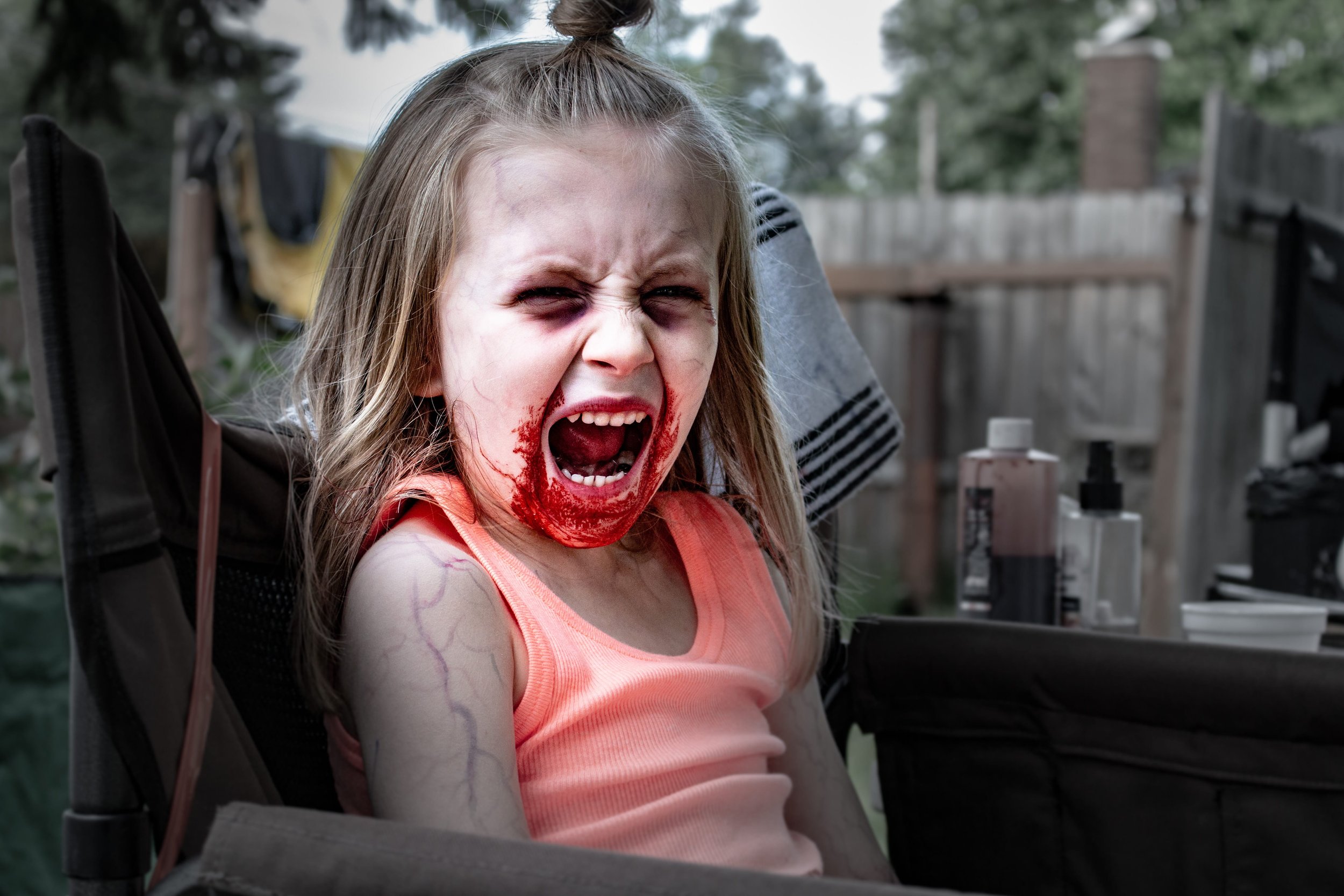A young girl with long blonde hair in a high ponytail, wearing a peach tank top, sitting in a stroller outdoors. She has blood on her chin and mouth, her face is contorted as if screaming or crying, and her arm has visible veins or scars. The backgro
