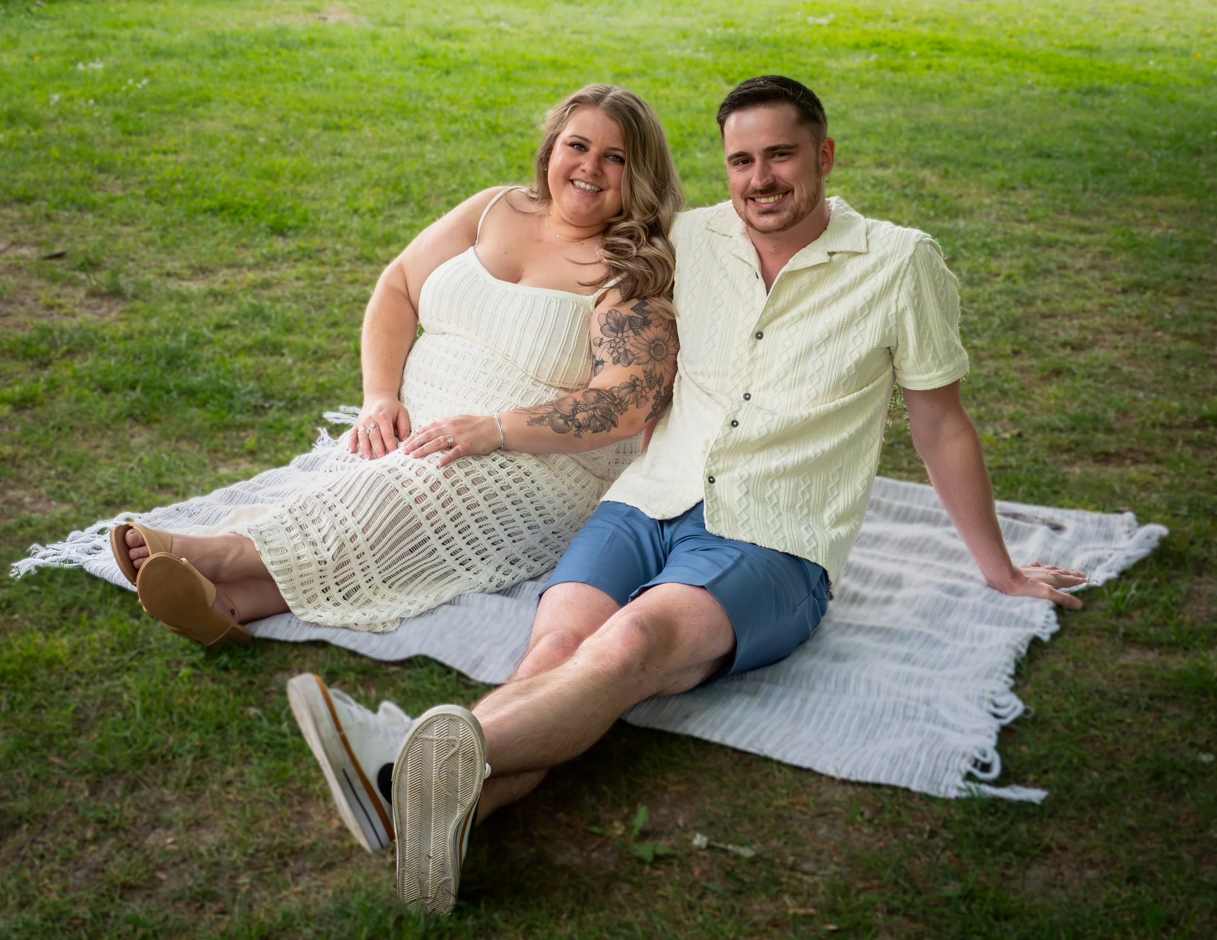 A smiling couple sitting on a blanket in a green park, the woman wearing a white dress with floral tattoos on her arm and the man in a light yellow shirt and blue shorts.