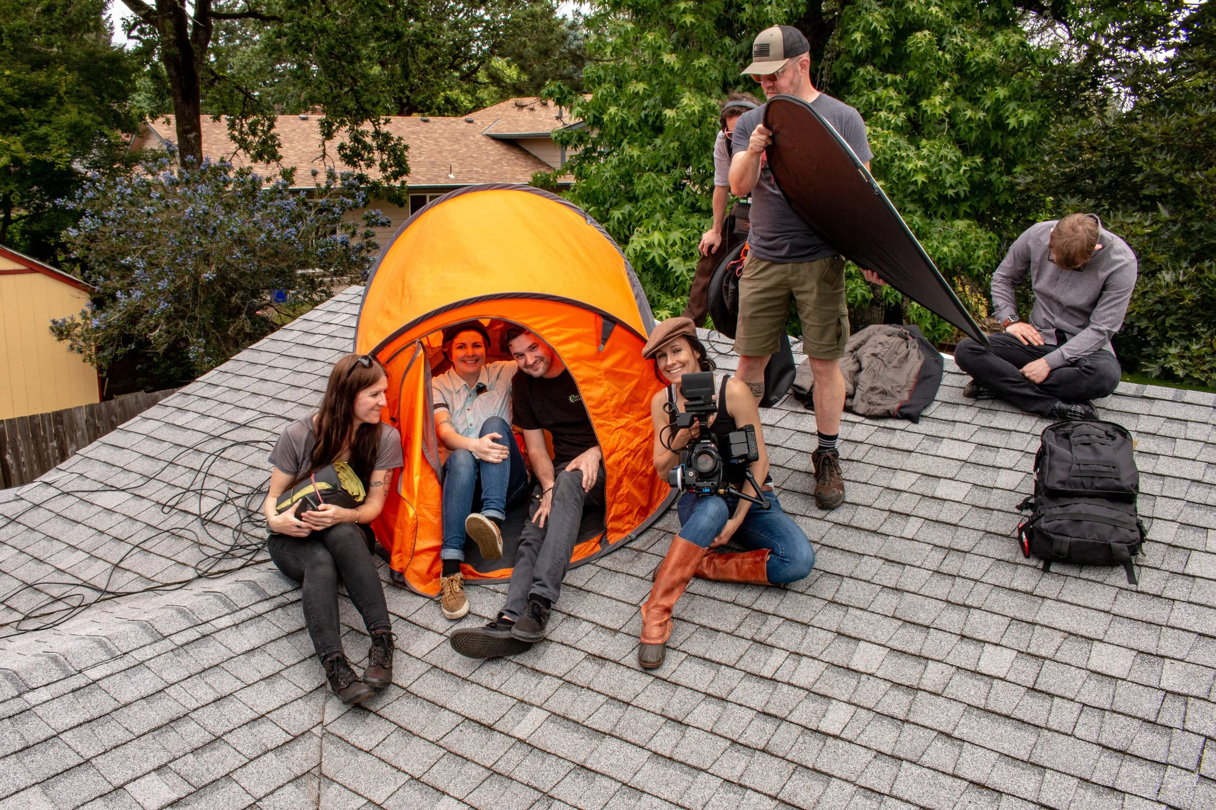 A group of six people on a rooftop with a small orange tent, some sitting inside and others standing around, preparing for an outdoor shoot on a cloudy day.
