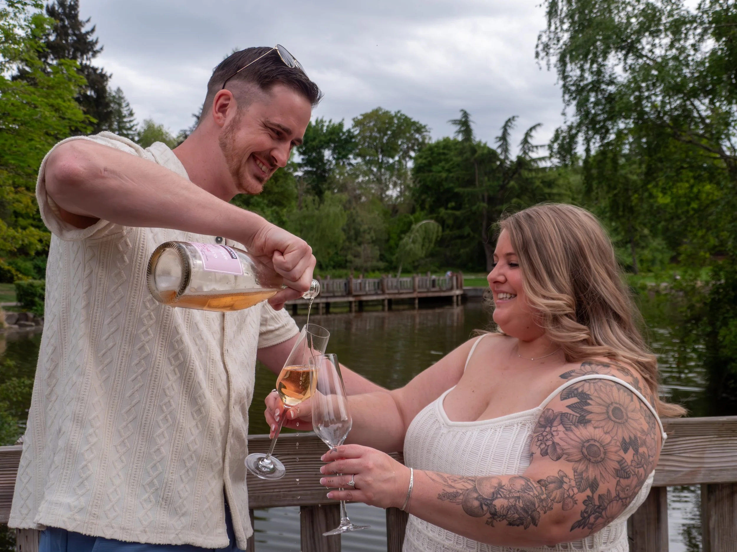 A man is pouring rosé wine from a bottle into a woman's champagne flute on a wooden dock beside a lake, surrounded by trees.