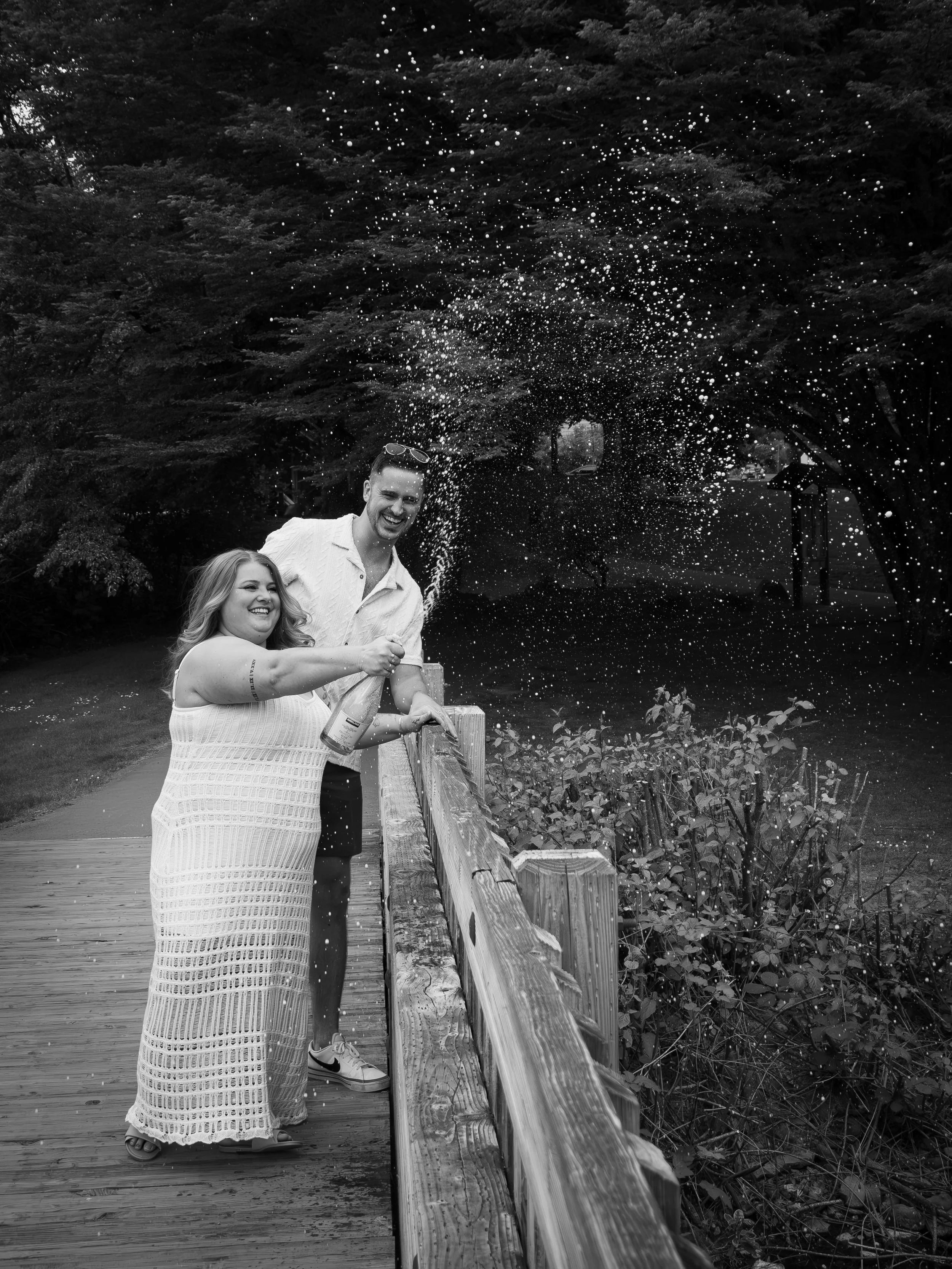 A black-and-white photo of a man and woman celebrating on a wooden bridge, with the woman spraying champagne and both smiling, surrounded by trees.