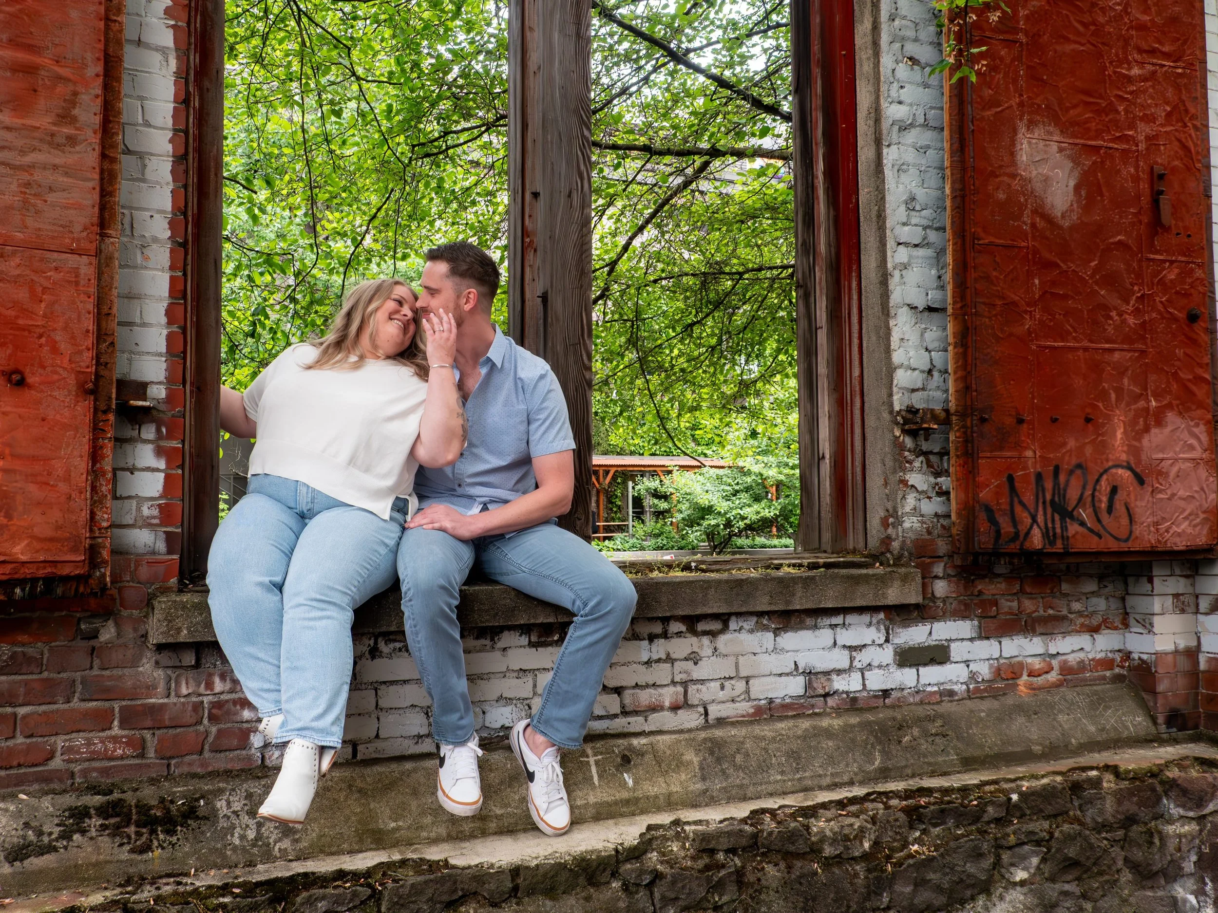 A couple sitting on a concrete ledge in front of a large open window with red wooden shutters, smiling and touching noses, with green trees and a building visible in the background.
