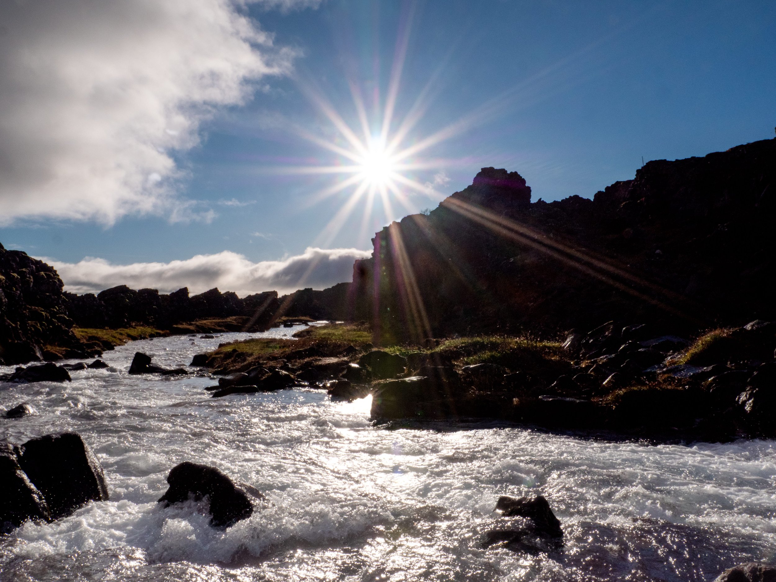 A scenic landscape of a river flowing through rocky terrain under a bright sun with rays, off-white clouds in a blue sky, and distant mountains.