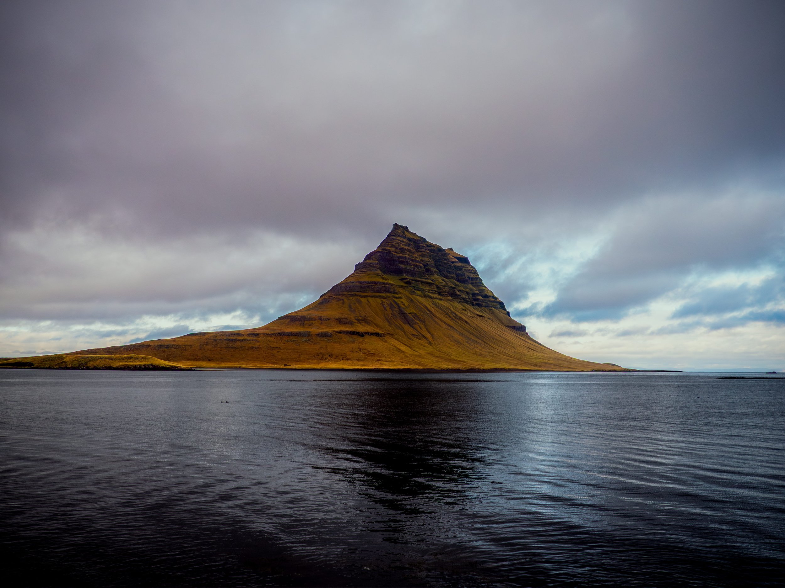 A mountain with a distinct conical shape rises from the sea, under a cloudy sky, with dark water in the foreground.