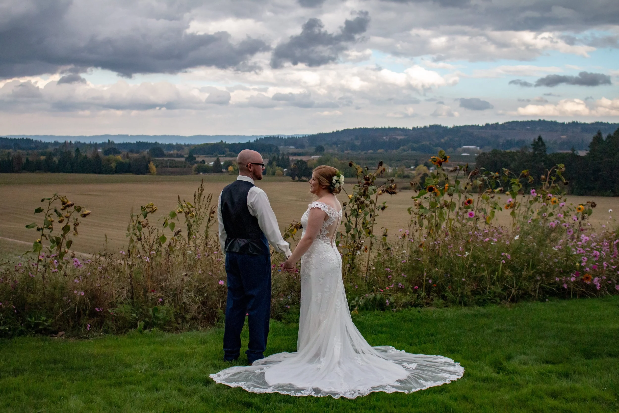 A bride and groom holding hands and facing each other outdoors, with a field and cloudy sky in the background.