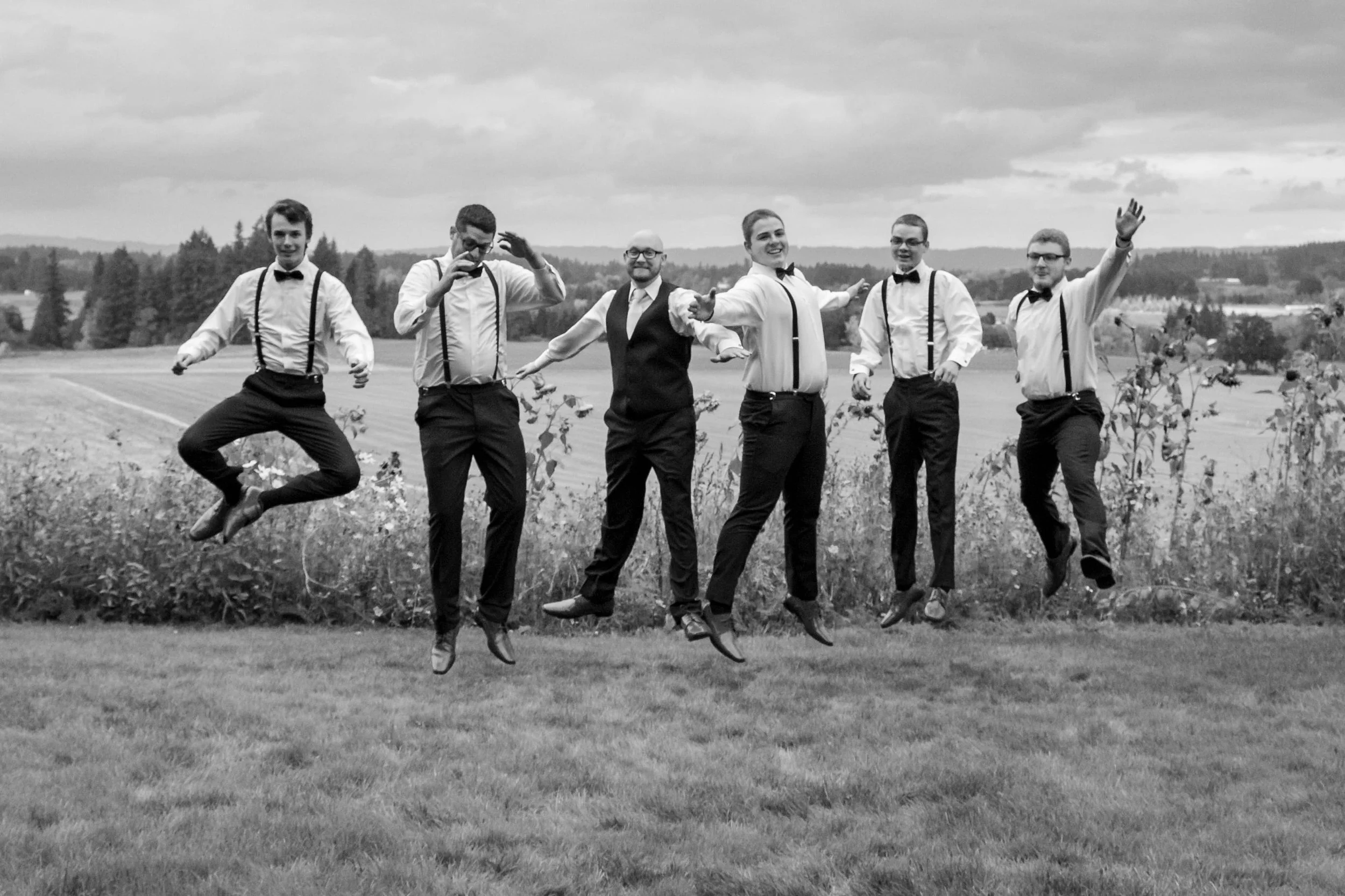 Group of six men in formal attire jumping in the air outdoors on grass with open field and trees in the background, black and white photo.