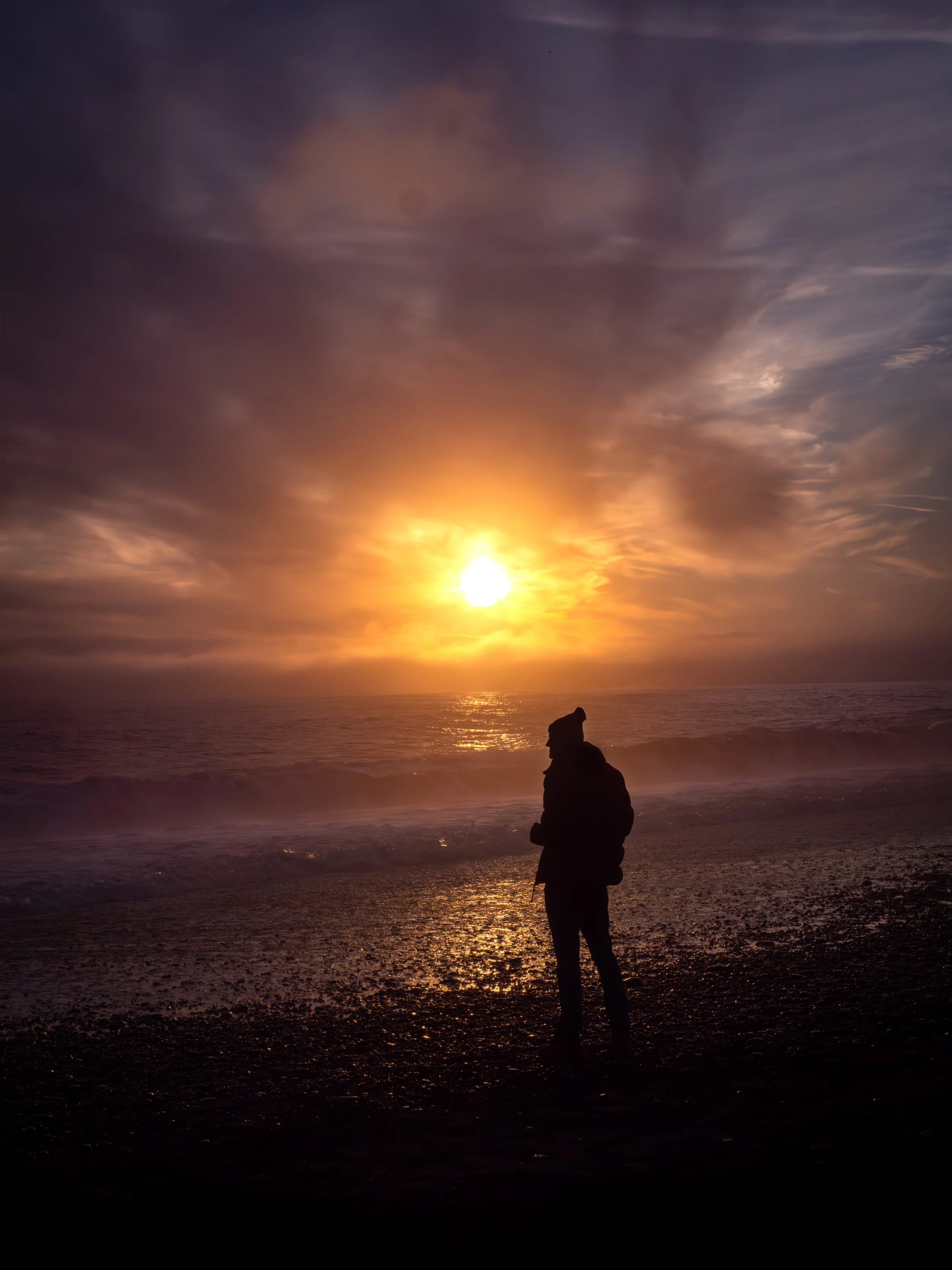 A person standing on a beach during sunset, silhouetted against the colorful sky with clouds and the ocean waves.
