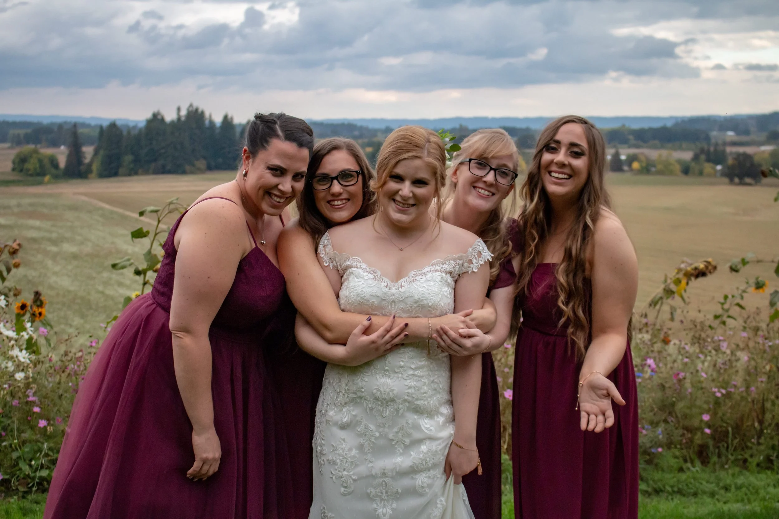 A bride in a white lace wedding dress is smiling while standing with four bridesmaids in burgundy dresses outdoors on a cloudy day with fields and trees in the background.