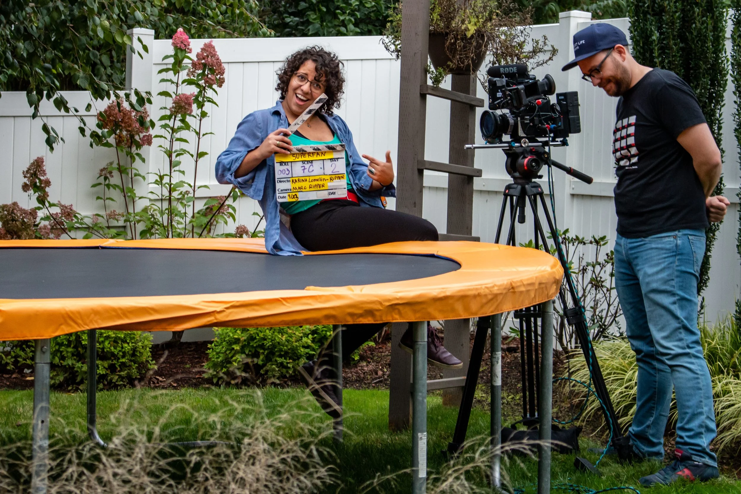 A woman sitting on a trampoline, holding a clapperboard, and smiling at the camera. A man operating a professional camera is standing nearby, in a backyard with plants and a white fence.