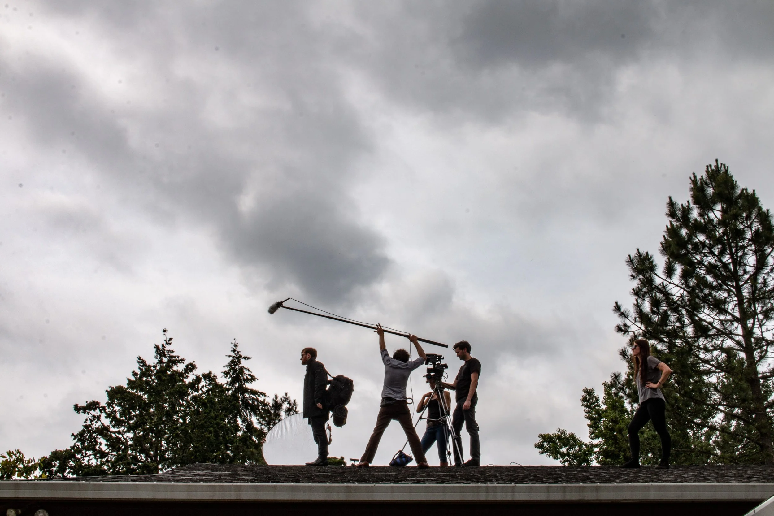 Film crew working on rooftop, with one person holding a boom microphone, another operating a camera, and others preparing equipment, under a cloudy sky.