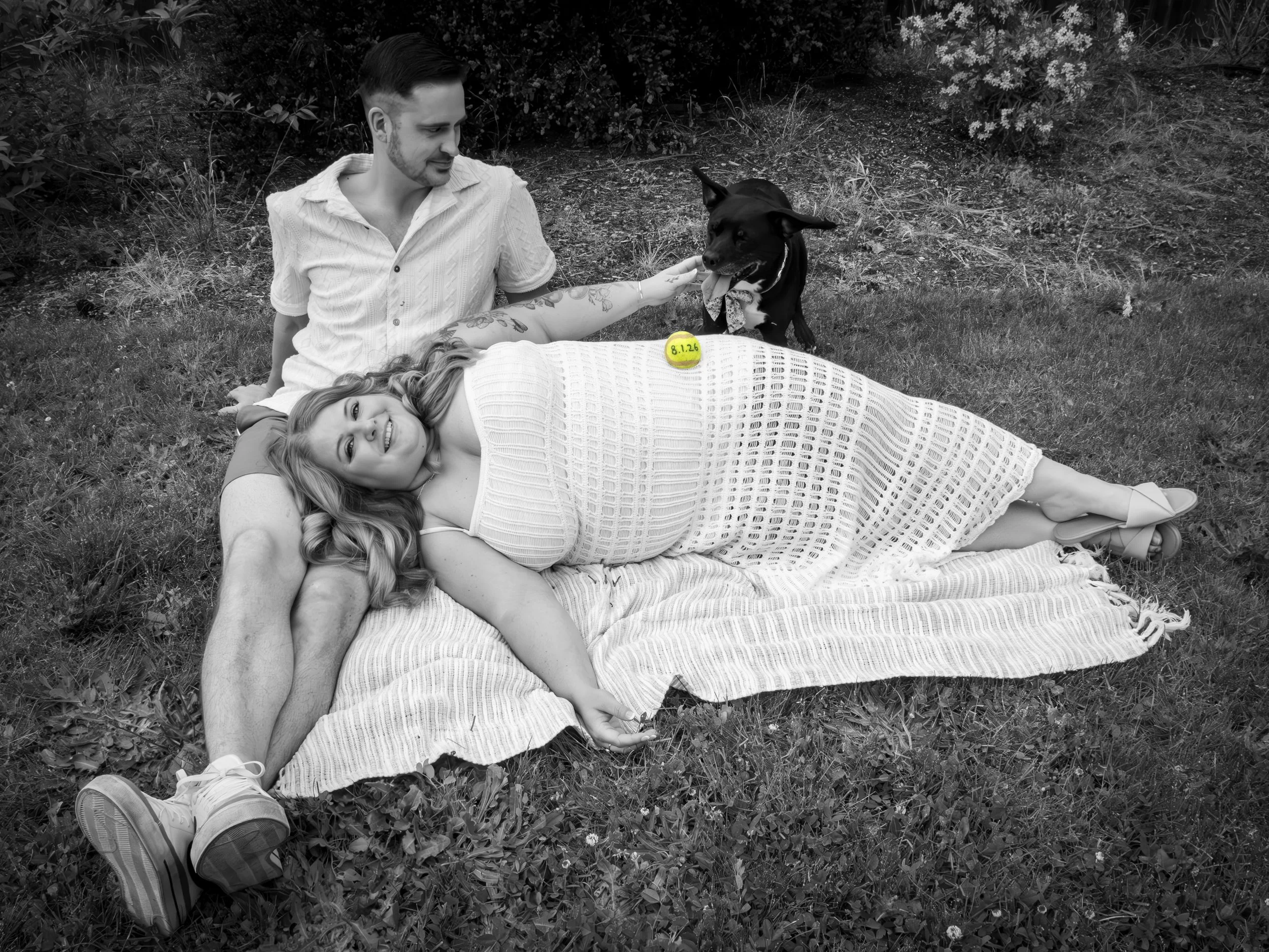 A pregnant woman lying on a blanket outdoors, smiling at the camera, with a man and a dog nearby. The man is sitting behind her, and the dog is sitting next to her with its tongue out. There is a yellow sticker on her belly with the date '8.1.26'.