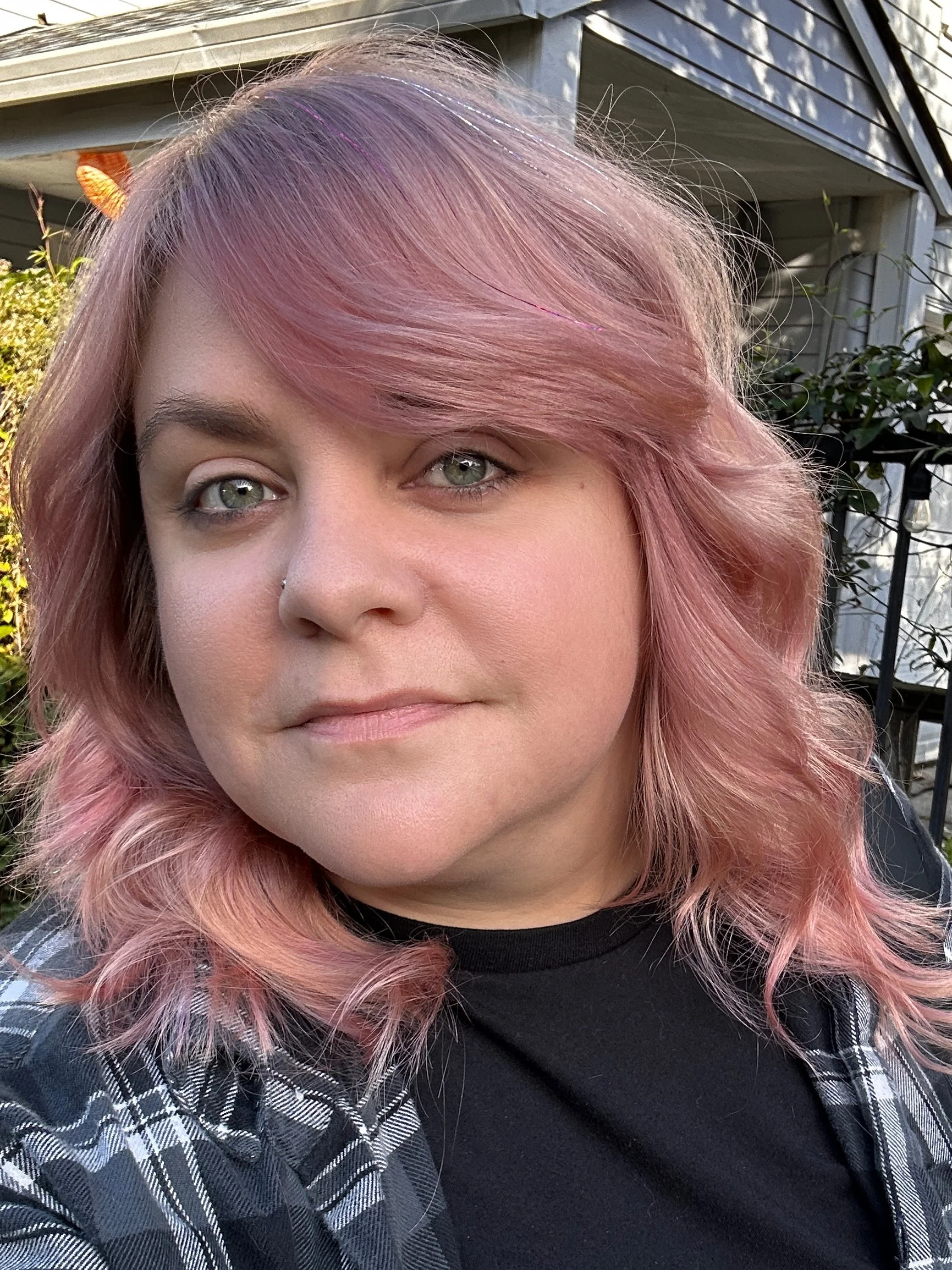 A woman with pink hair, wearing a black shirt and a black and white plaid shirt, standing outdoors near a house with siding and a garden.