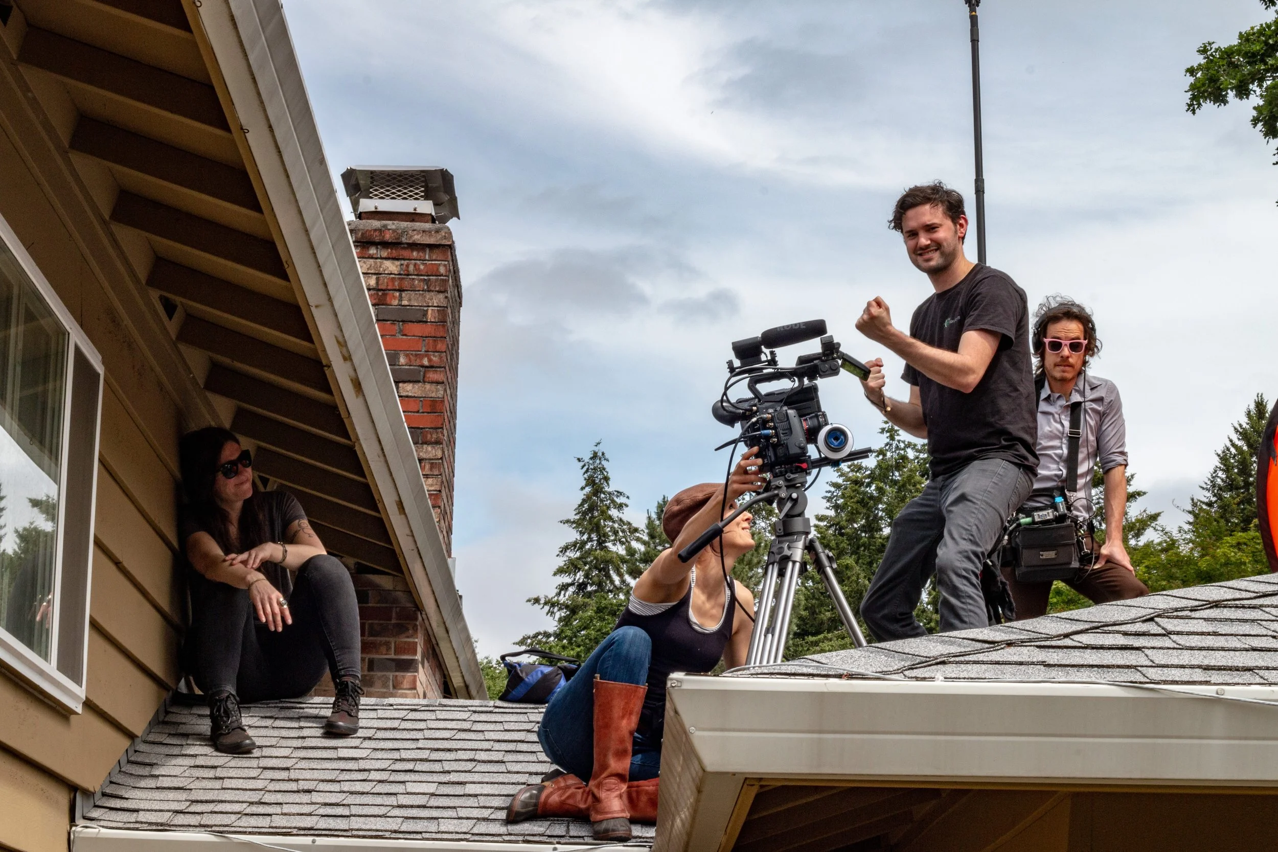 A film crew on a rooftop, with one person sitting by a window on the side of a house, another operating a camera on a tripod, and a third person standing and celebrating, while a woman kneeling and adjusting equipment.