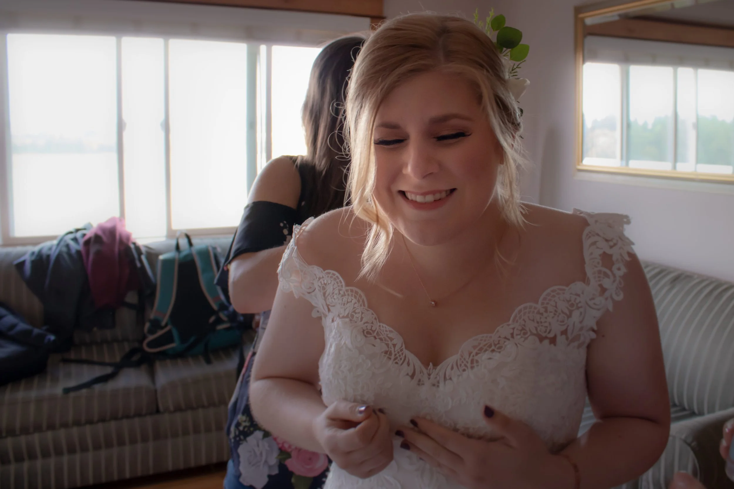 A woman wearing a wedding dress with lace details is smiling with her eyes closed, as someone helps her with her dress in a bright room with large windows and a sofa with clothes and bags on it.