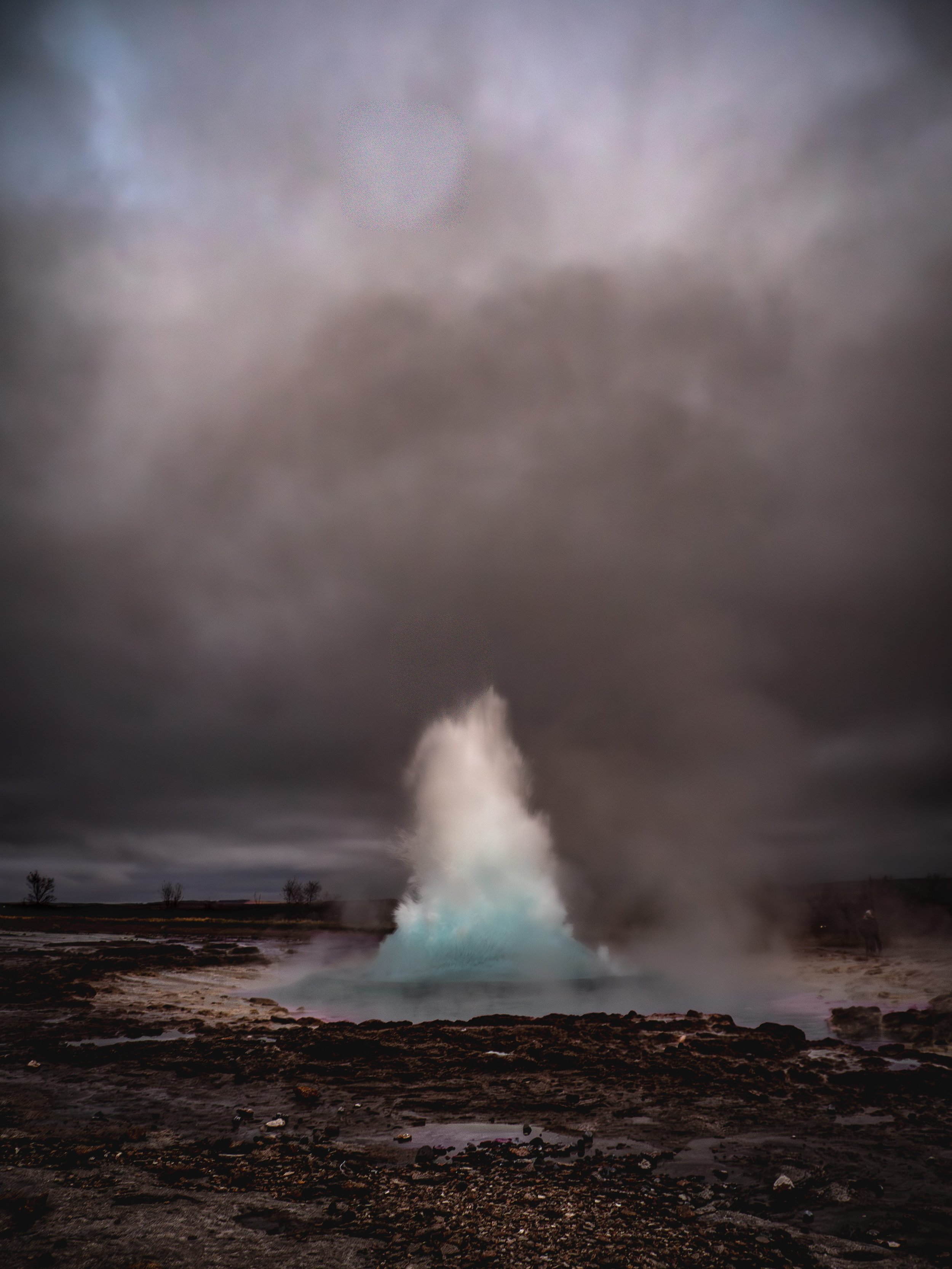 A geyser erupting in a geothermal area with rocky ground and dark, cloudy sky overhead.