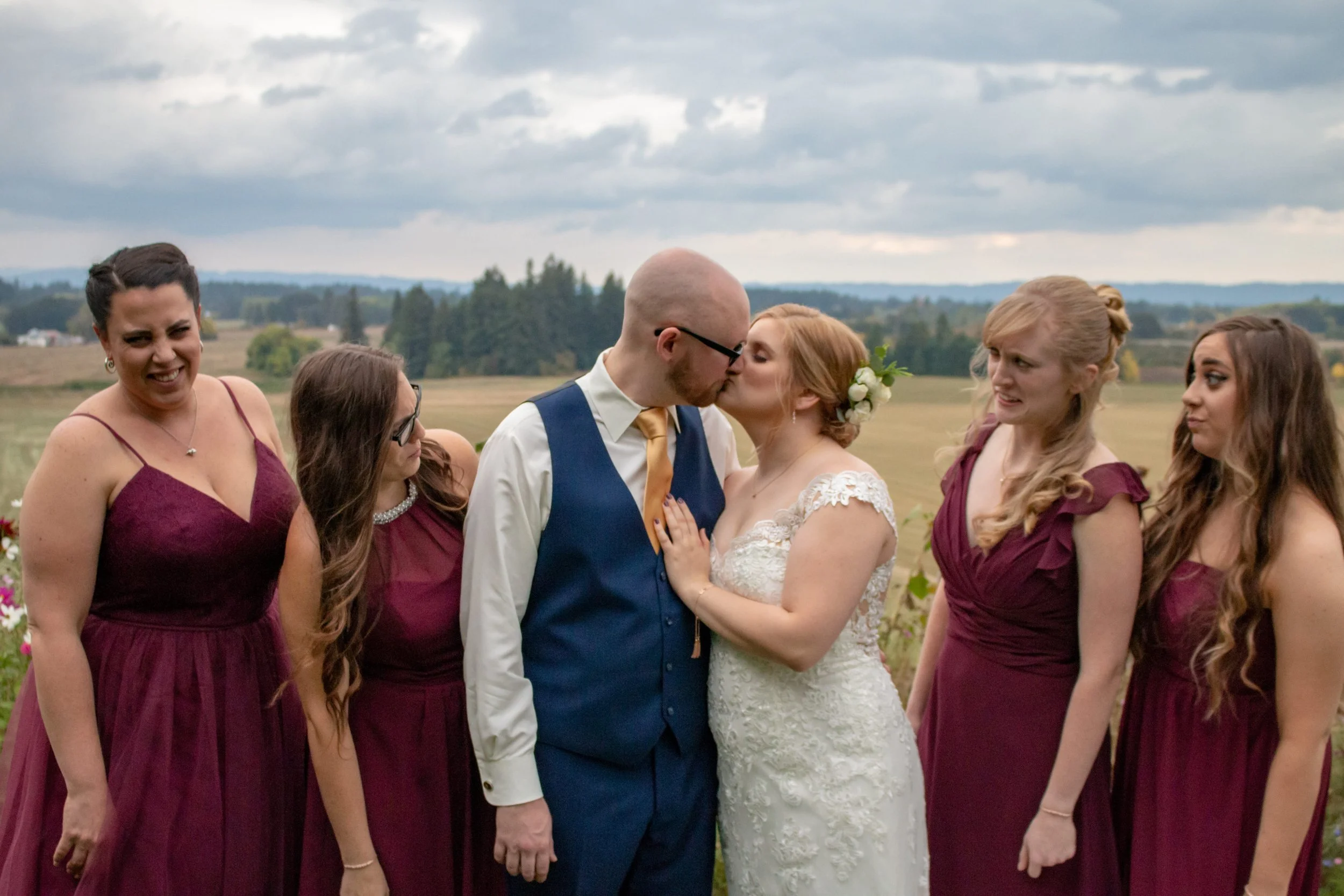 A wedding scene outdoors with the bride and groom sharing a kiss, surrounded by four bridesmaids in burgundy dresses, while standing in an open field with trees and cloudy sky in the background.