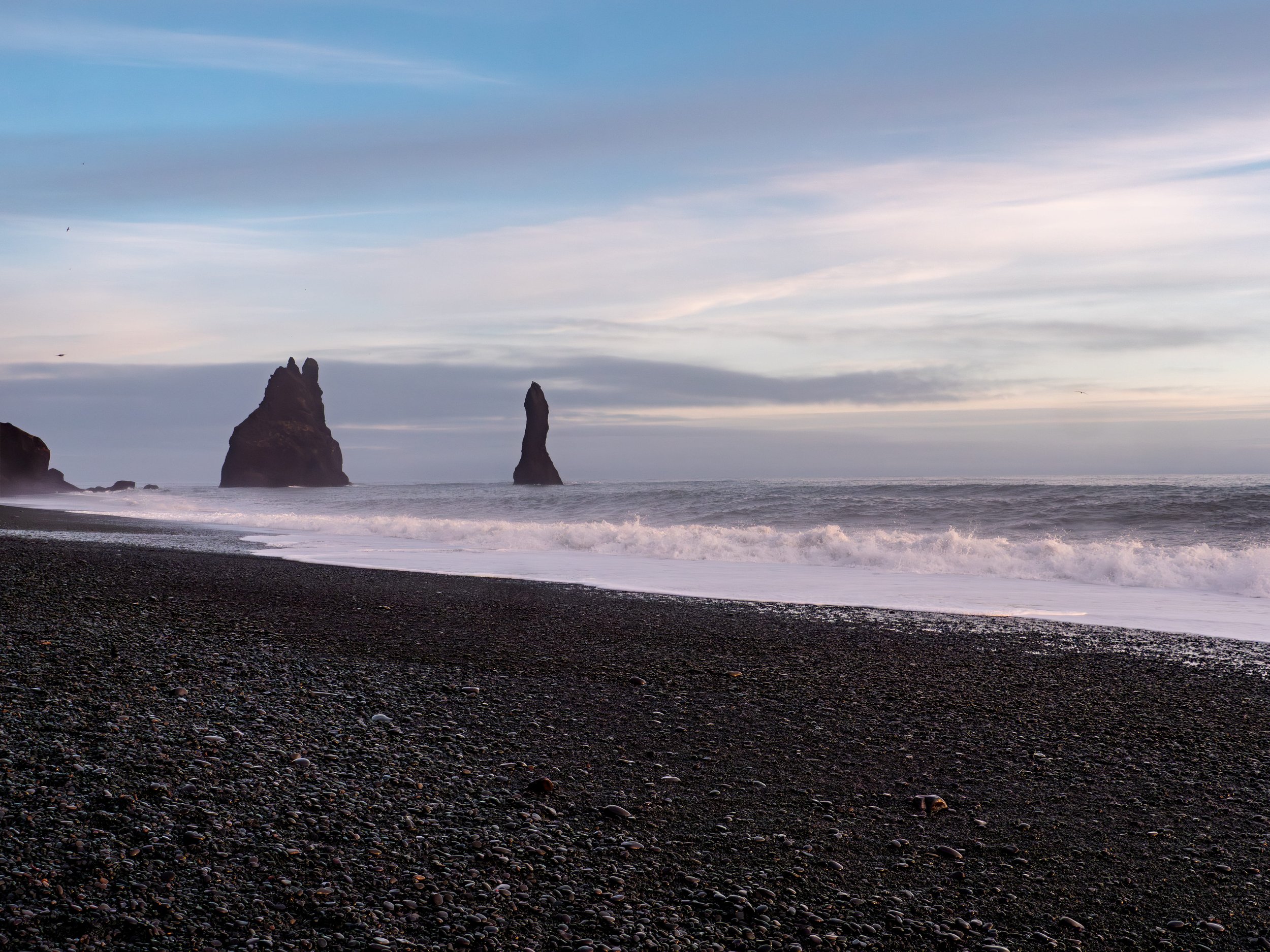 A black sand beach with two tall rock formations in the ocean under a cloudy sky.