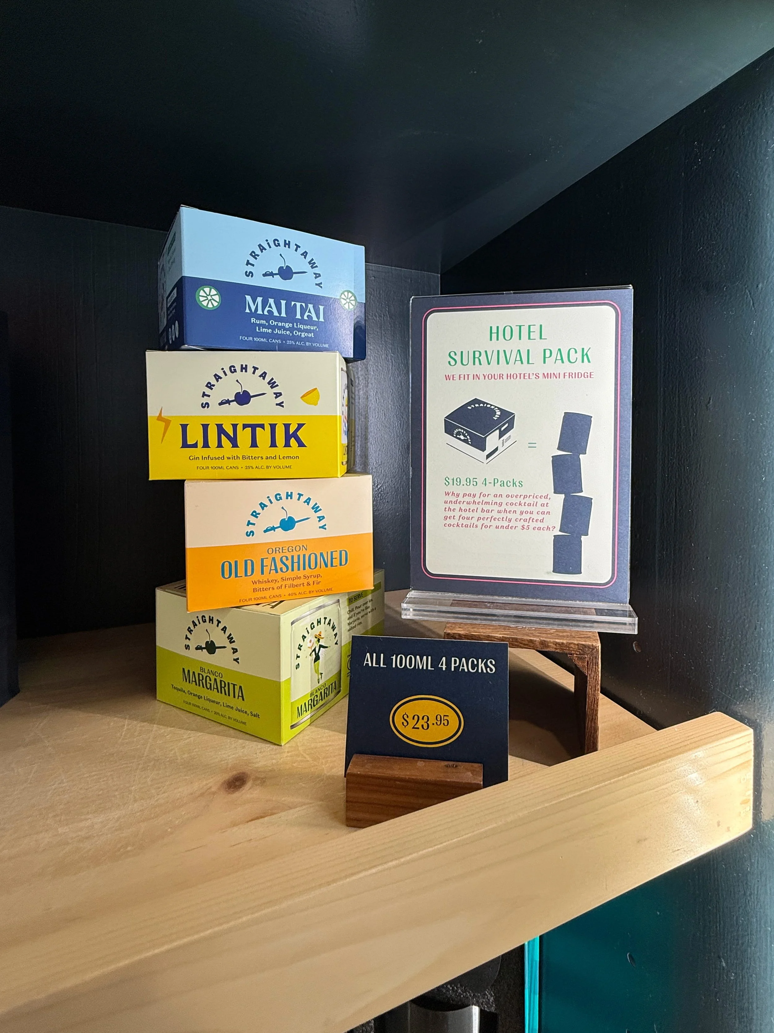 Display of four boxes of cocktail mixers on a wooden shelf, with a sign offering four 100ml packs for $23.95.