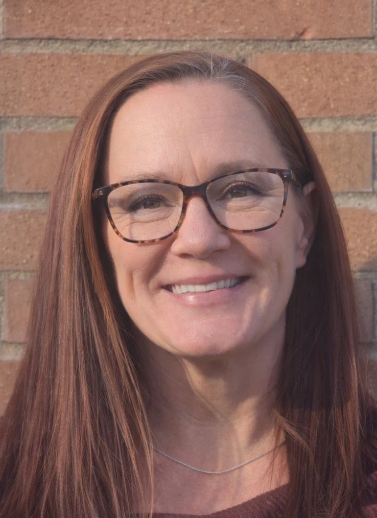 A woman with long reddish-brown hair wearing tortoiseshell glasses, smiling, standing in front of a brick wall.