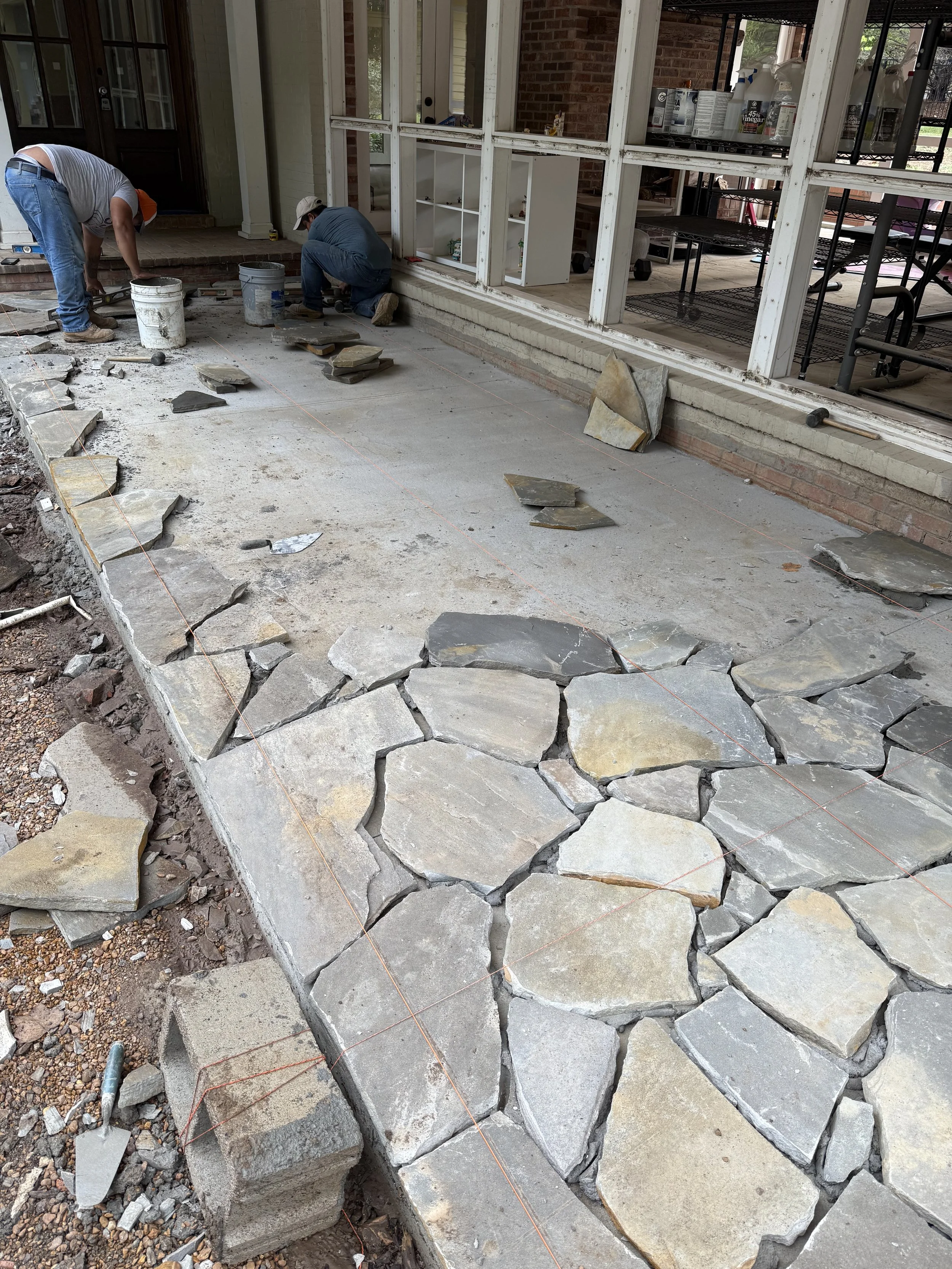 Two workers installing flagstone on a concrete patio outside a house, with construction tools and materials around.