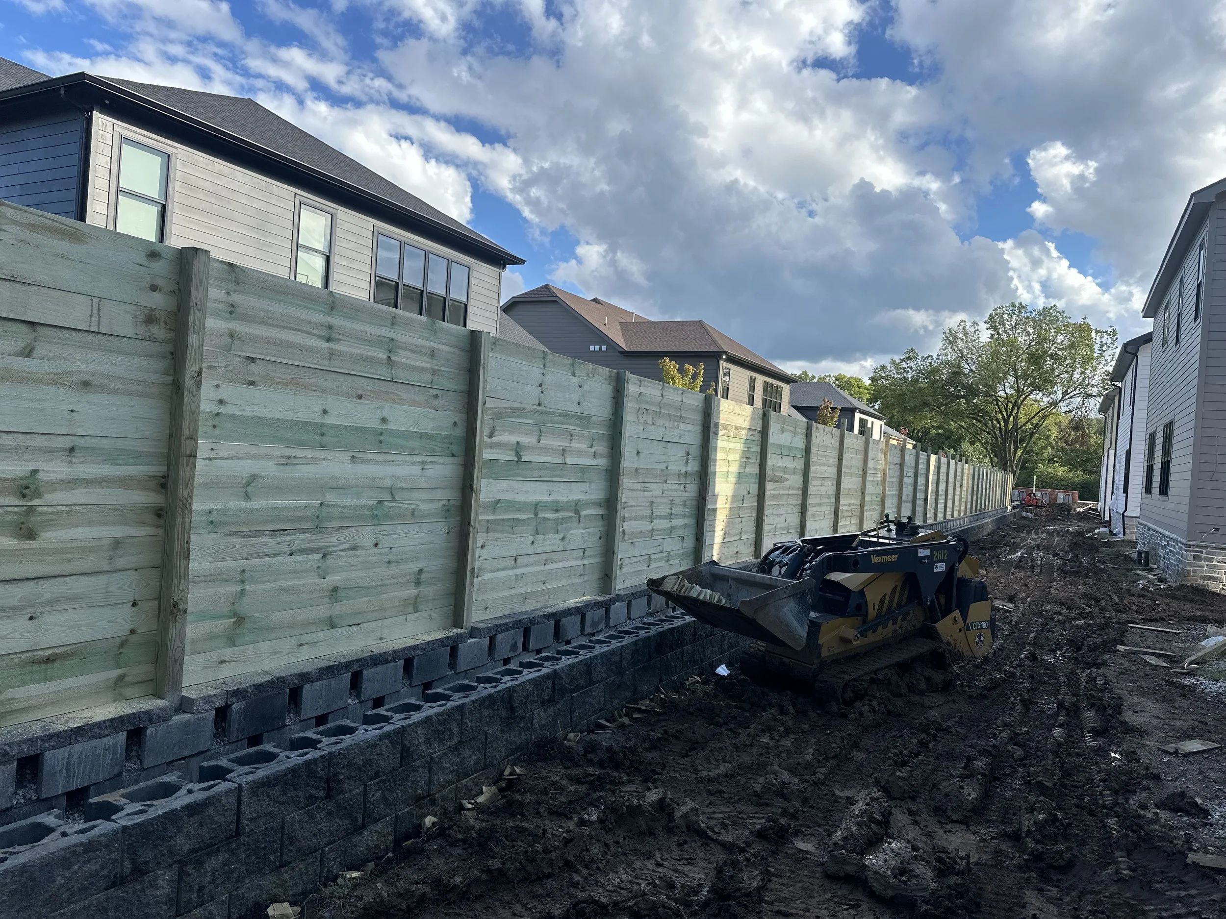 A construction site with a new wooden fence being built along a dirt pathway between residential houses. A small construction vehicle is on the dirt, and the sky is partly cloudy with sunlight shining through.