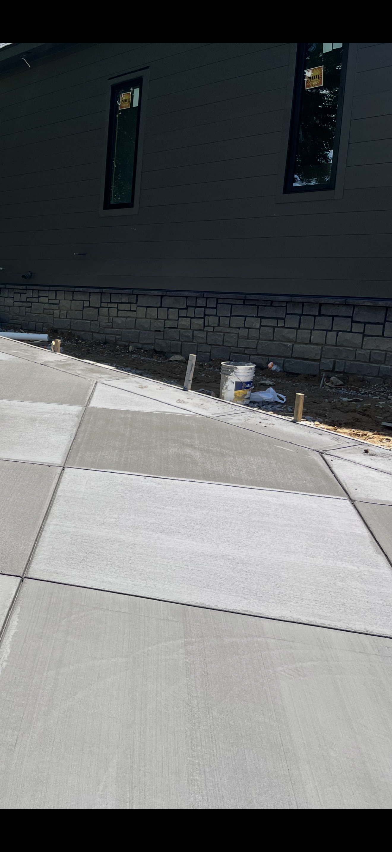 Exterior of a building under construction with a concrete sidewalk in the foreground, construction materials, and uneven dirt ground along the base of the building.