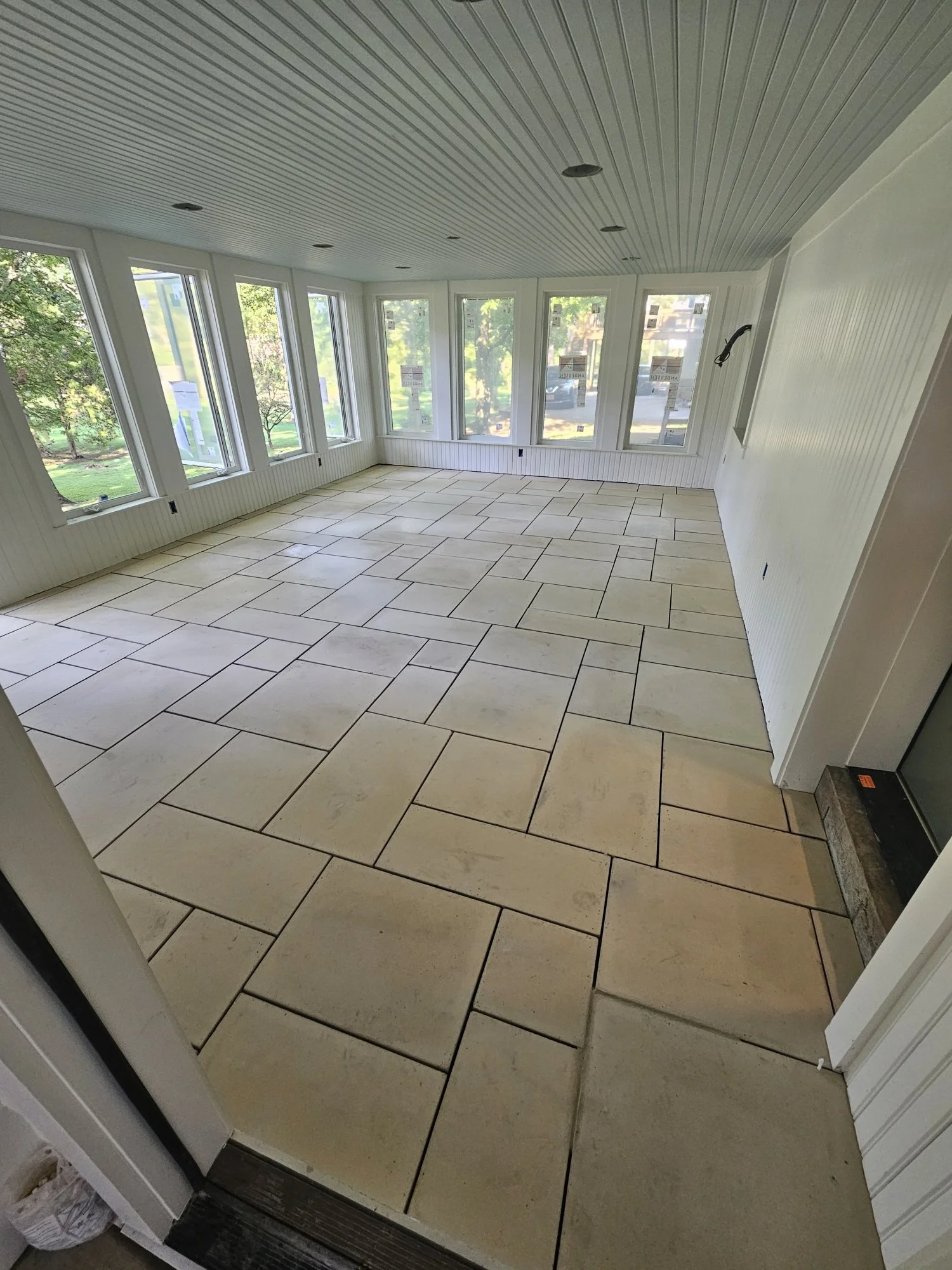 Empty sunroom with large windows, beige tile flooring, and white beadboard walls and ceiling.