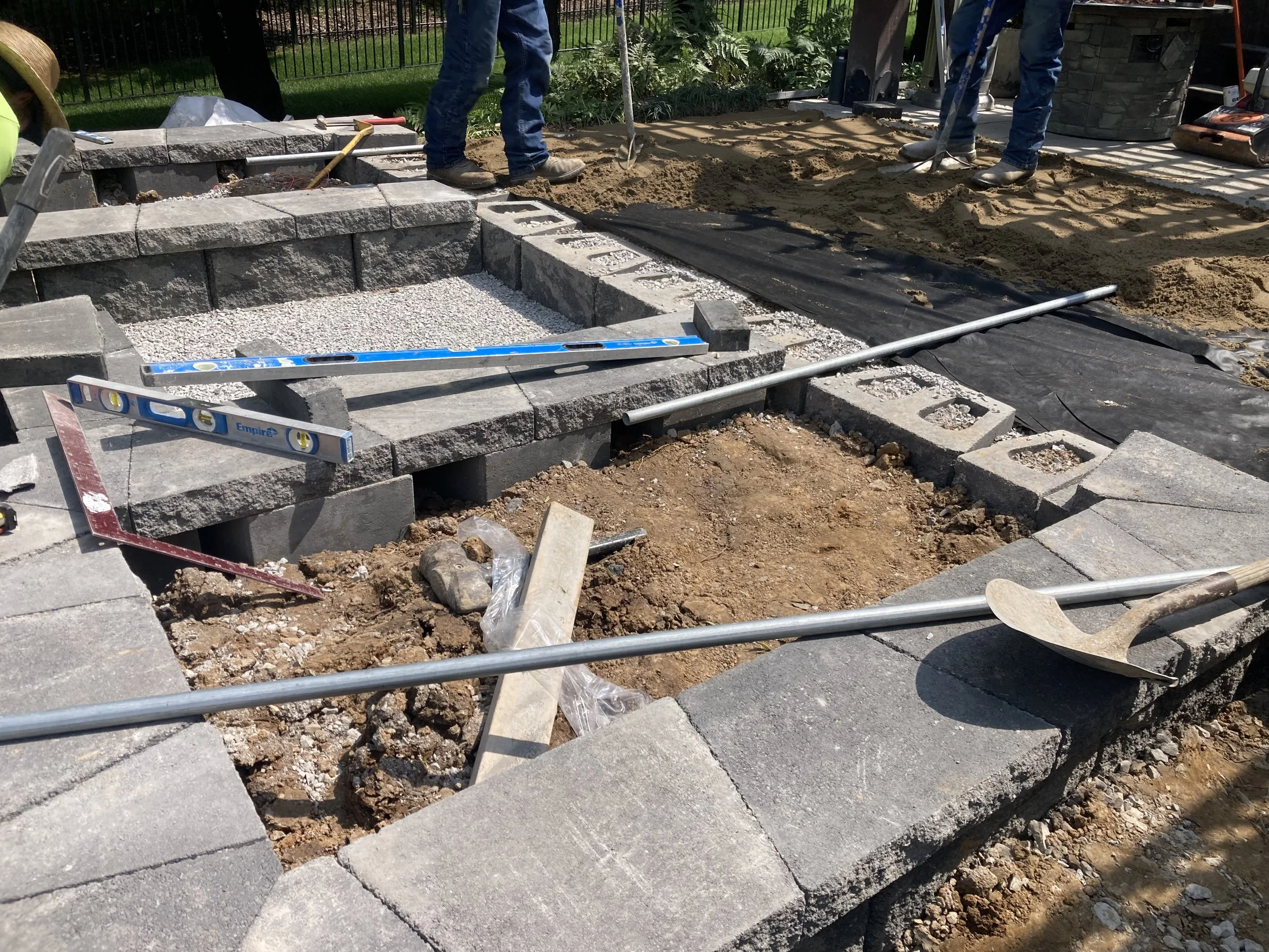 Construction workers building a stone patio or pathway with large gray pavers, using level tools, shovels, and measuring equipment on a sunny day.