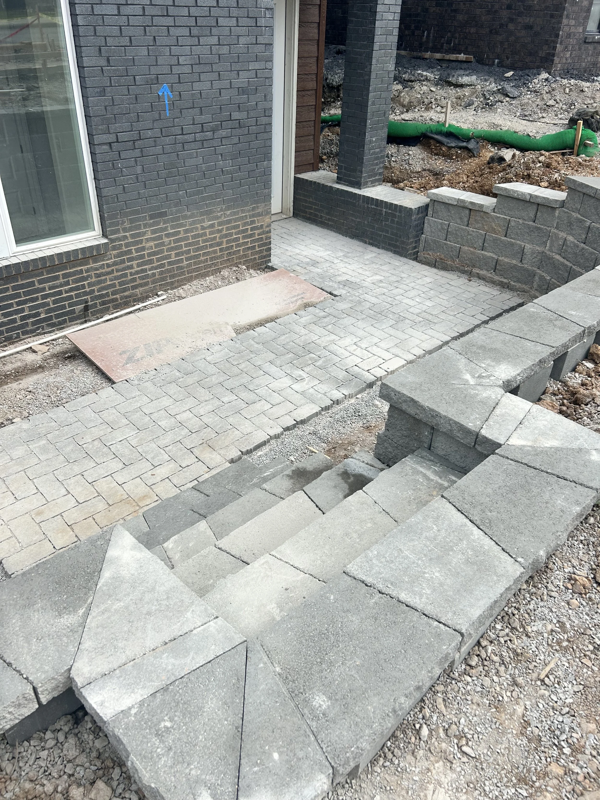 Construction site showing a partially completed brick and stone patio with stairs and walls, with exposed dirt and building materials around.