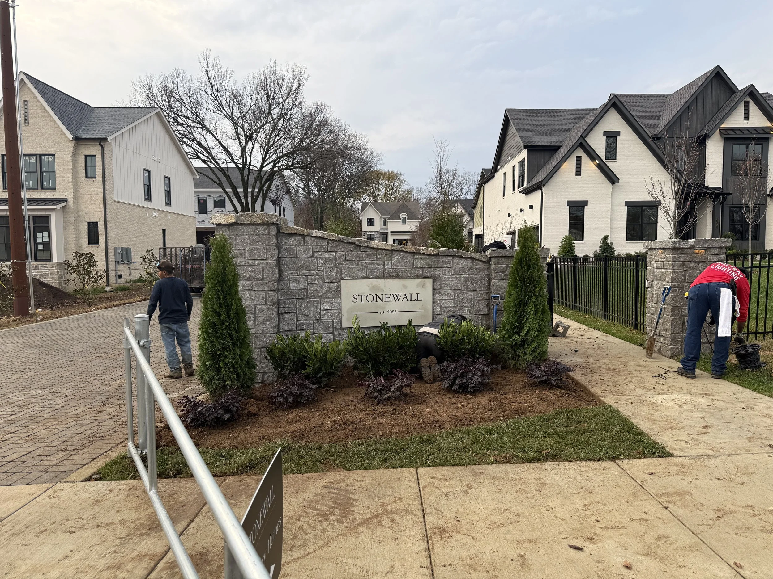 People working on landscaping in front of a stone wall entrance to a neighborhood named Stonewall, with modern houses in the background.