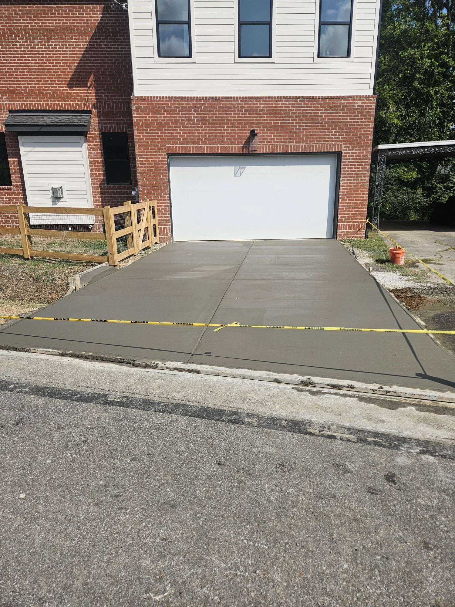 Newly poured concrete driveway in front of a white garage door, with caution tape around the area, adjacent to a brick and siding house.