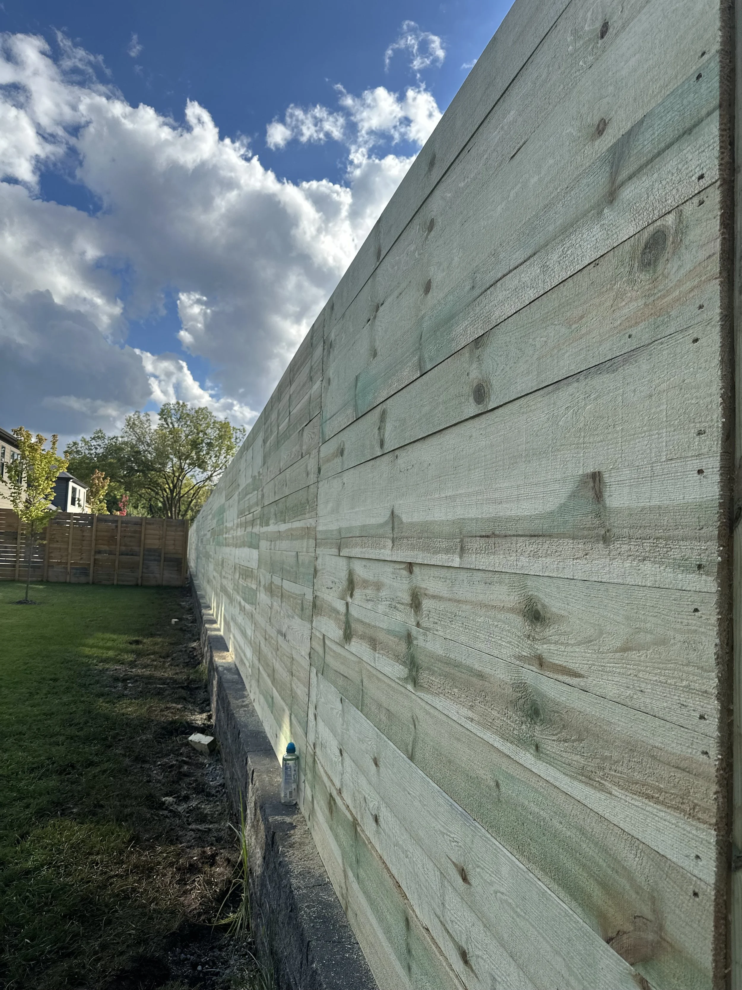 A wooden fence outdoors on a sunny day with partly cloudy sky, portion of a yard with grass, some trees, and neighboring houses in the background.