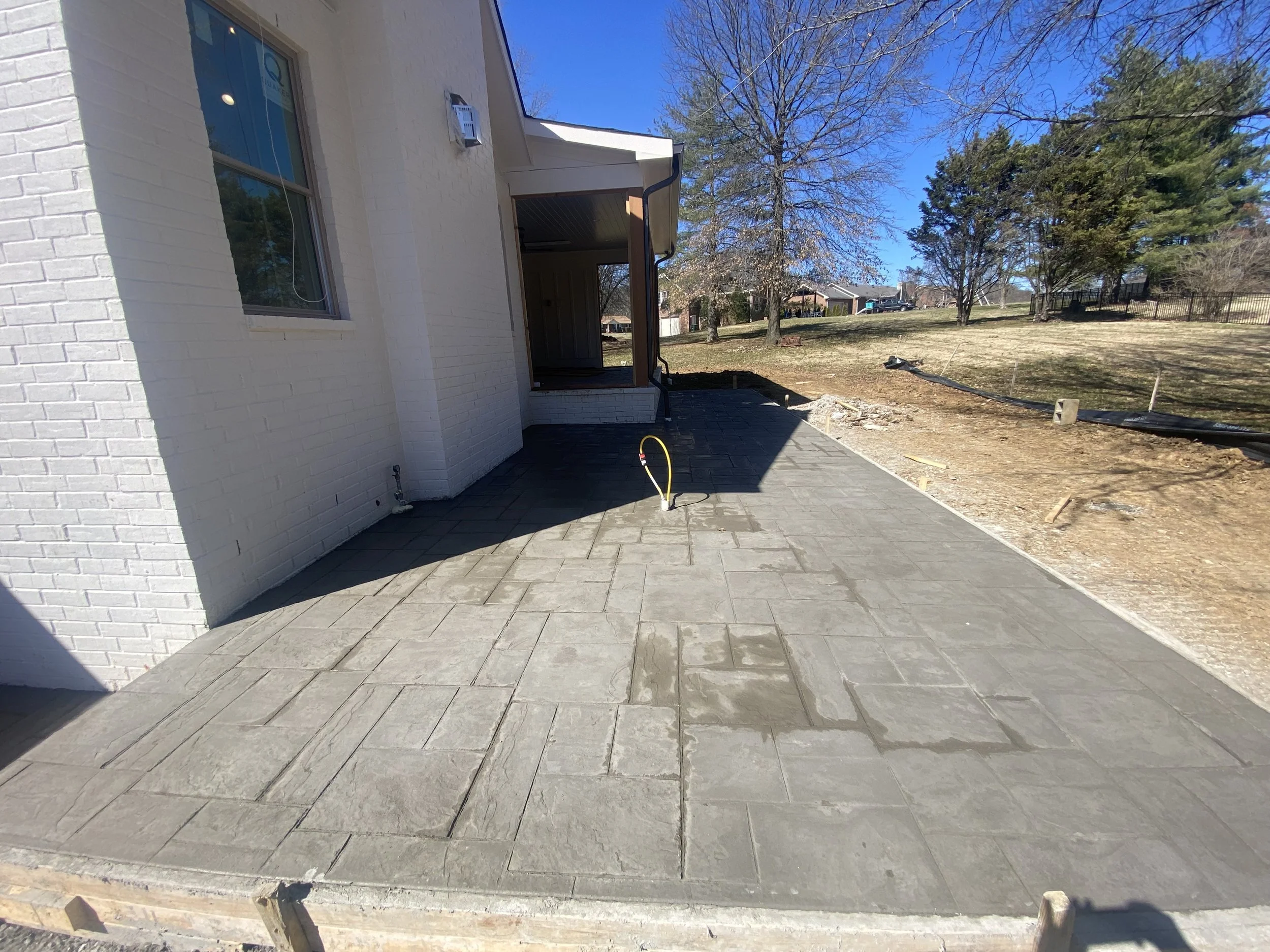 Newly installed gray concrete patio with a patterned surface in a backyard, adjacent to a white brick house, with trees and blue sky in the background.