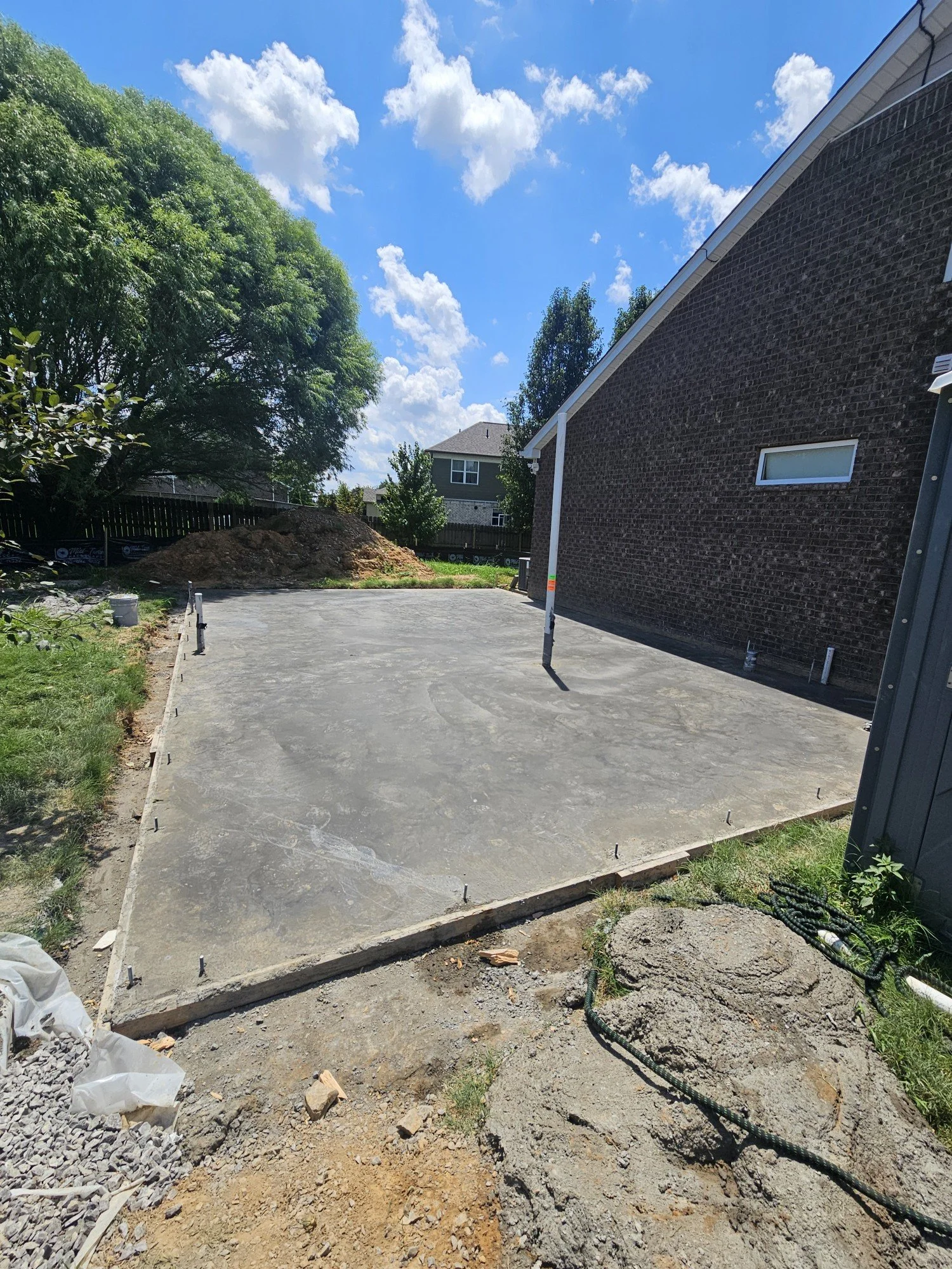 A freshly poured concrete slab next to a brick house, with construction stakes and a pile of dirt in the background, under a partly cloudy sky.