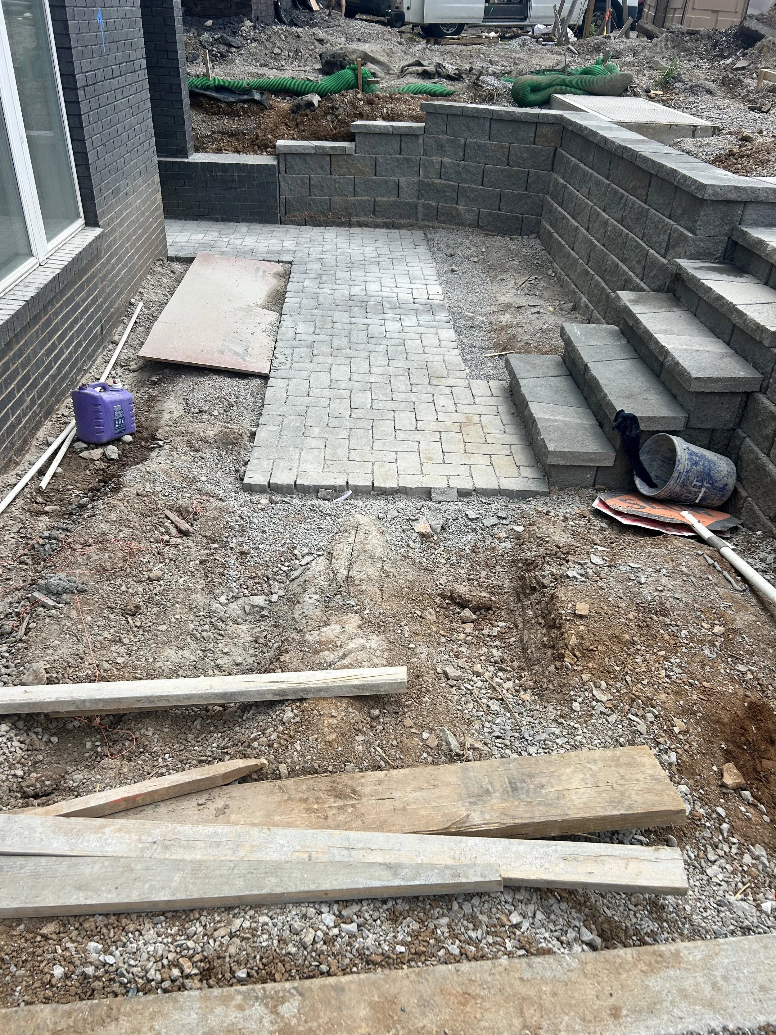 A construction site showing a newly installed brick patio, stone retaining wall, and stairs under construction. Various construction materials and tools are visible, with dirt and gravel in the foreground.