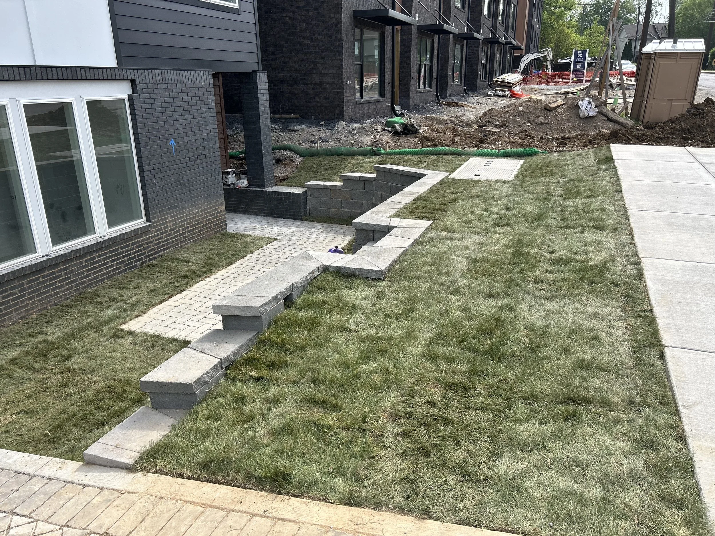 A landscaped backyard with a brick pathway, some grass, and a retaining wall under construction near a row of black brick houses.
