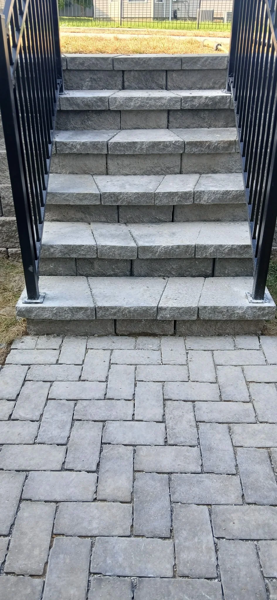 A set of outdoor stone stairs with black metal railings on both sides, leading up to a grassy yard with a fence and houses in the background. The stair treads are made of rectangular gray stone blocks.