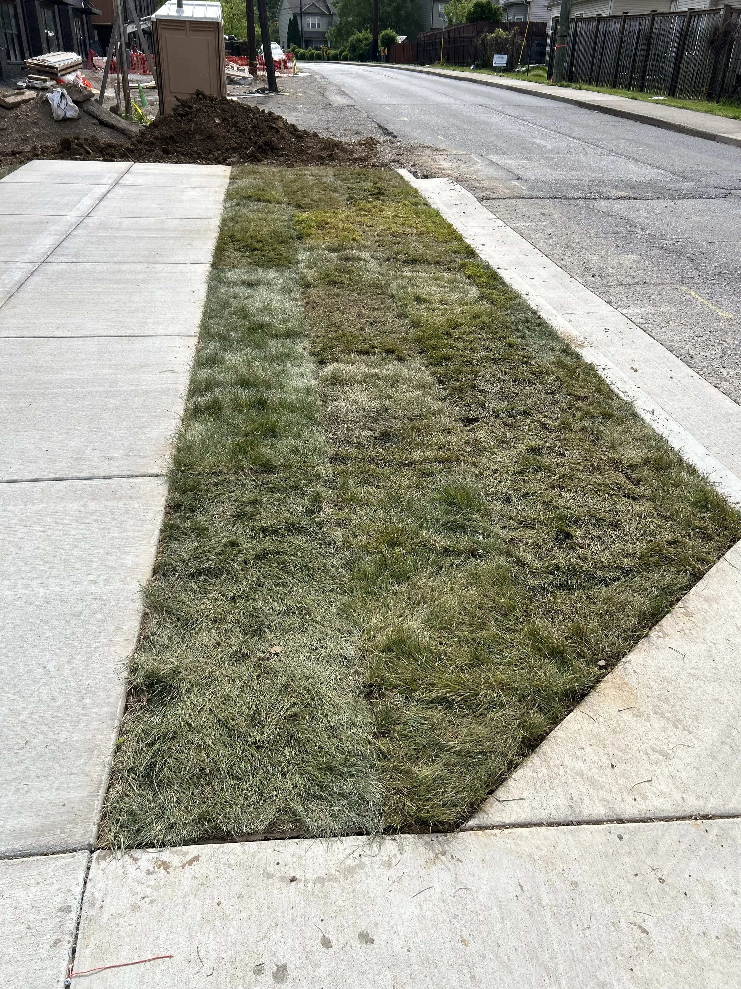 A sidewalk next to an outdoor construction site with patches of grass, a storage shed, and construction materials in the background.