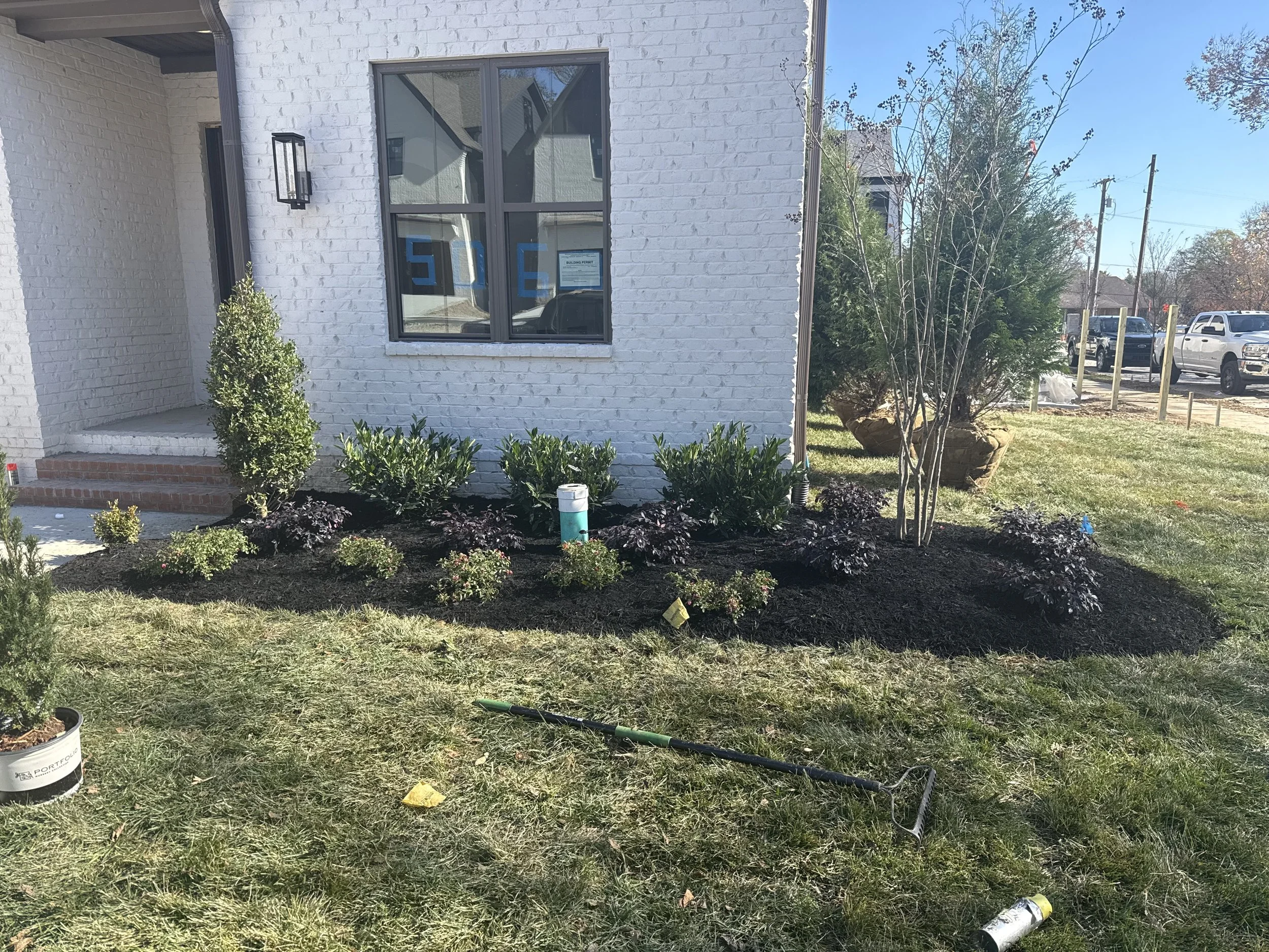 Freshly landscaped front yard with dark mulch, small shrubs, and a young tree in front of a white brick house. Construction tools and materials are on the grass, including a rake, spray can, and a small container.